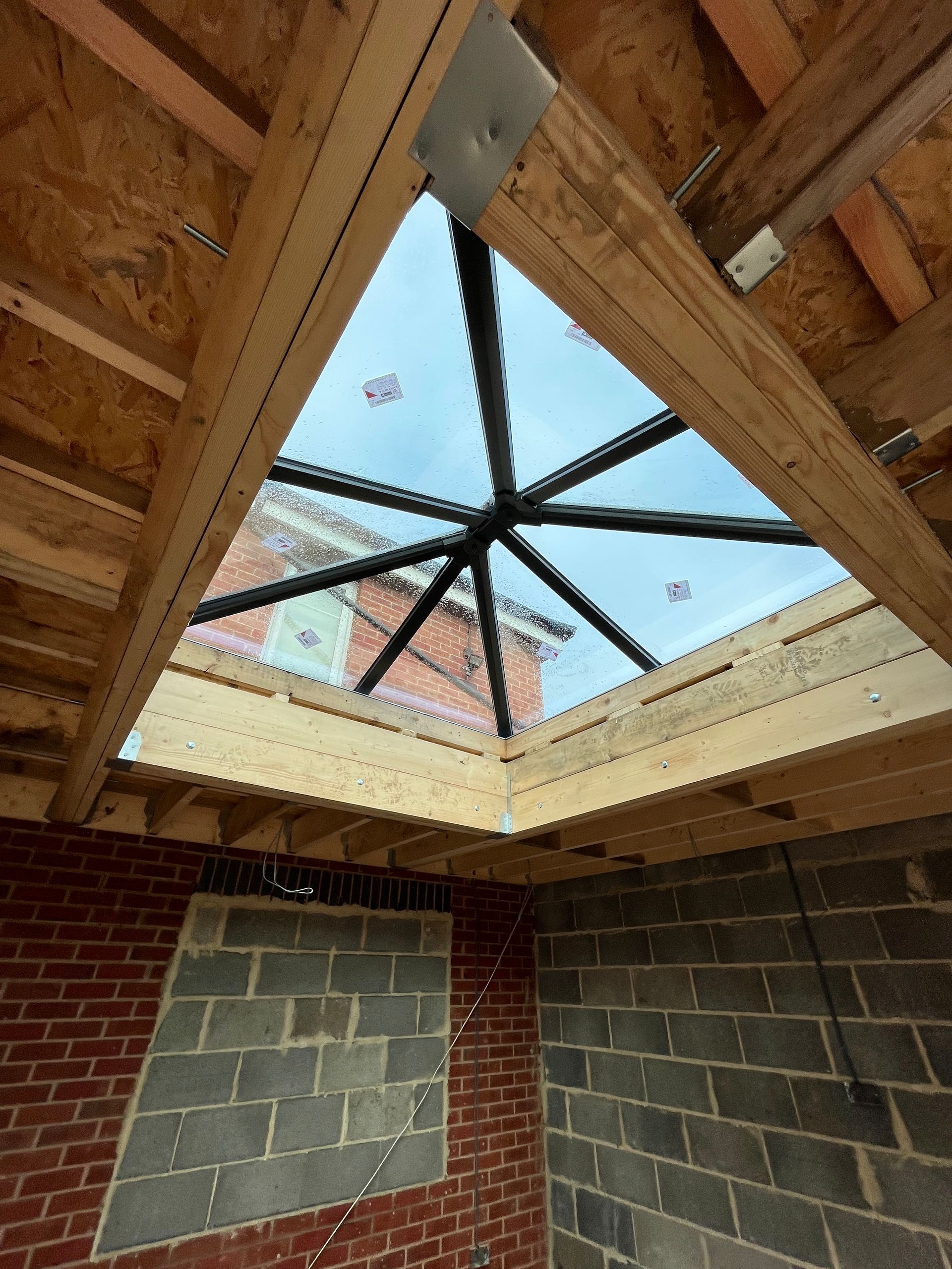 Interior view of a construction site with a triangular skylight in the roof. Wooden beams support a black-framed window.