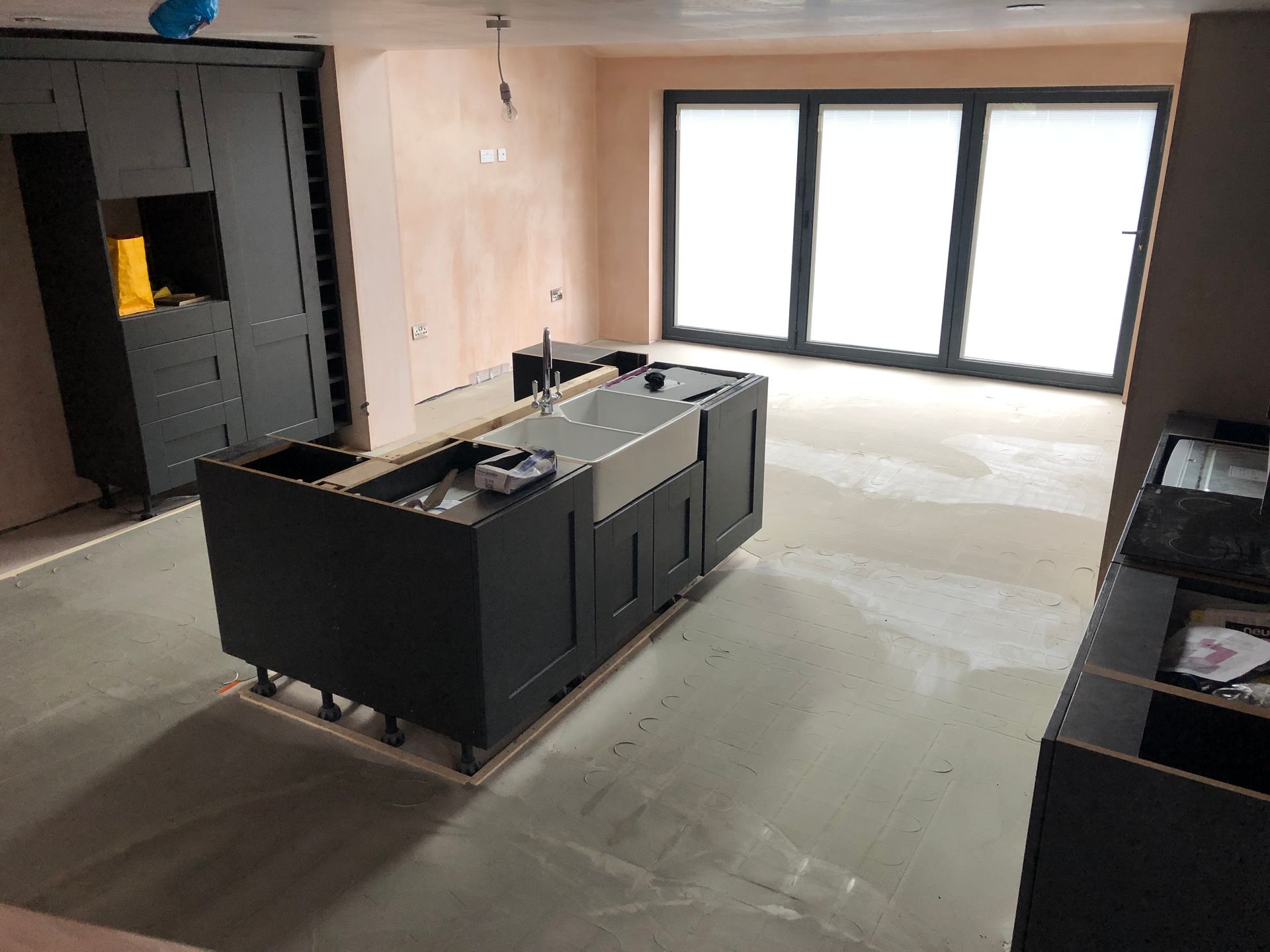 Kitchen under construction: dark gray cabinets, stainless steel sink, large windows, and exposed walls.