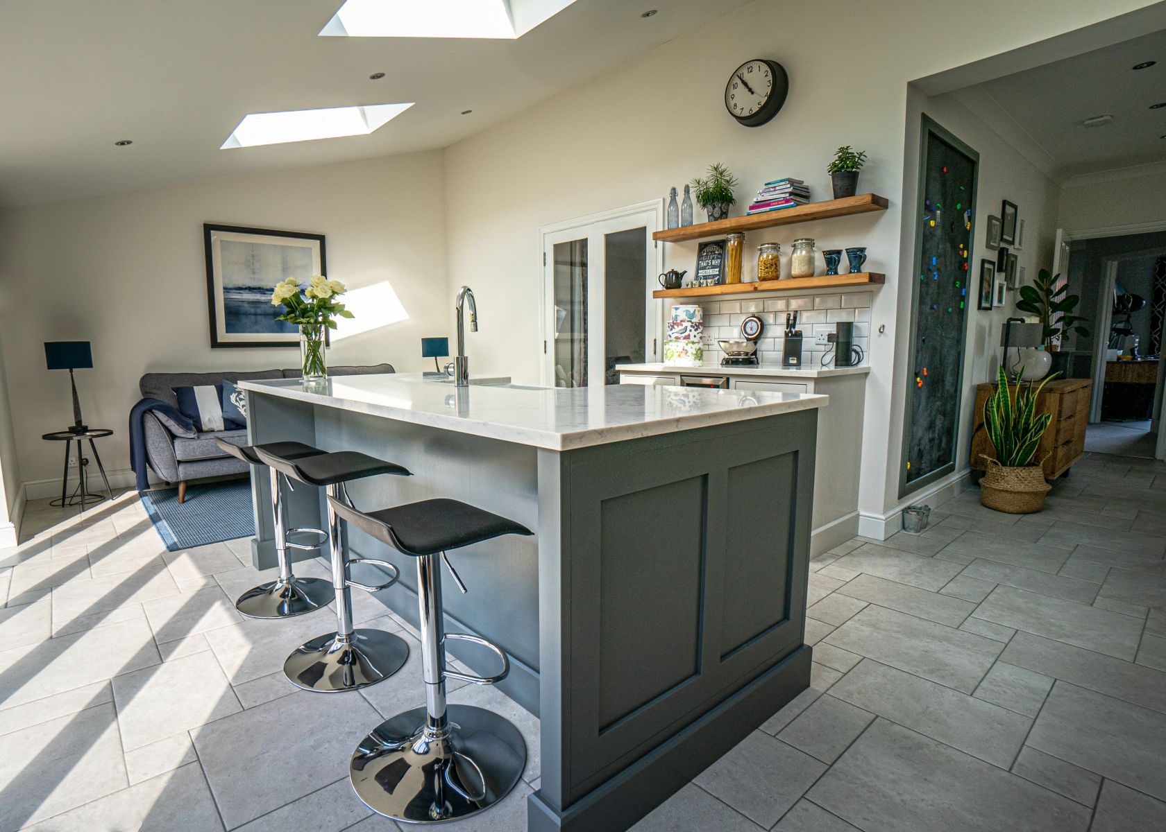 Modern kitchen with gray island, seating, and open shelves. Includes a couch, gray tiled floor, and natural light.