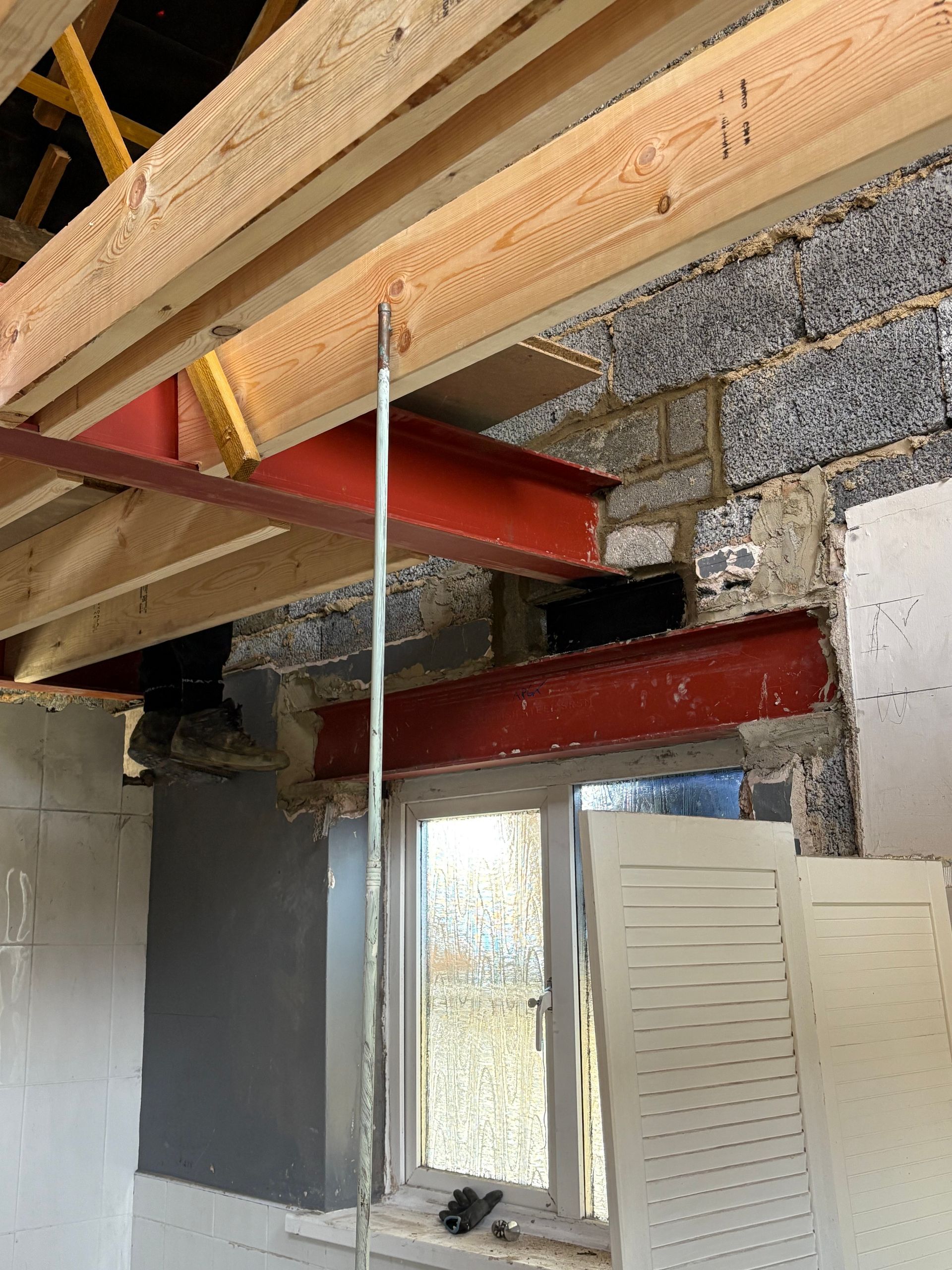 Interior view of a construction site with red steel beams supporting wooden beams and a window.