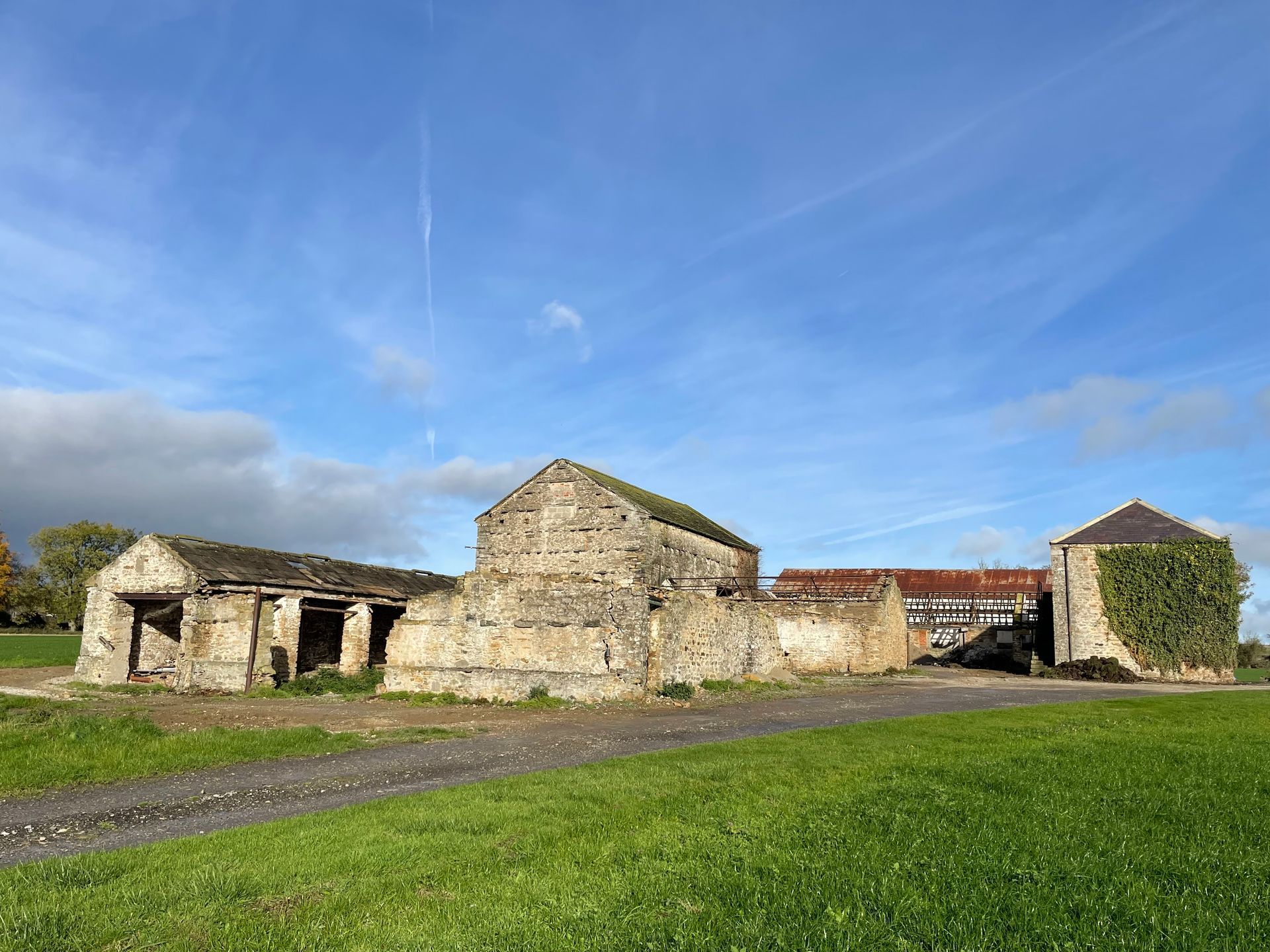 Old stone farm buildings under a blue sky, with green grass and a dirt road in the foreground.