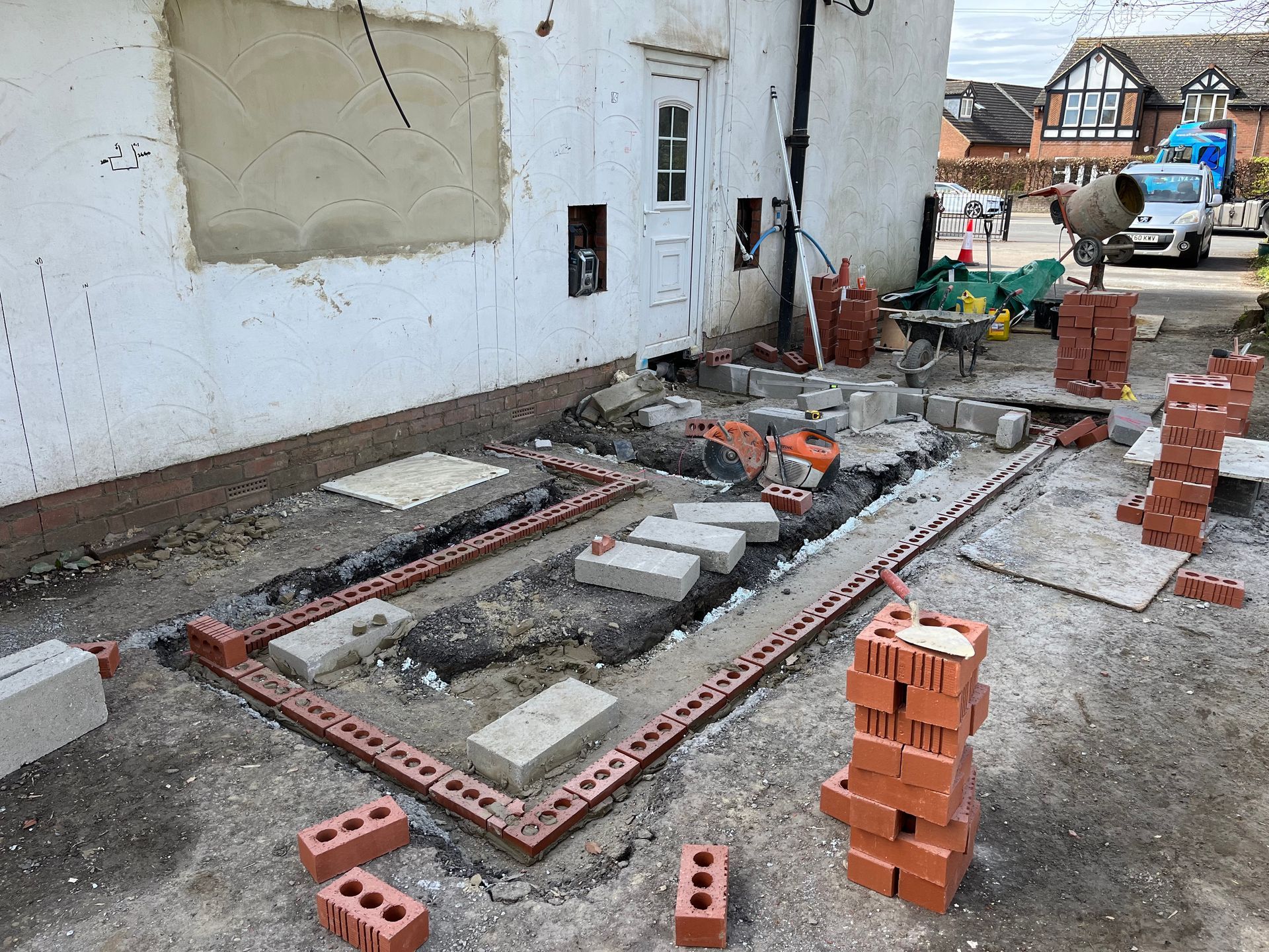 Construction site: foundation being laid with red brick and concrete blocks next to a white building.