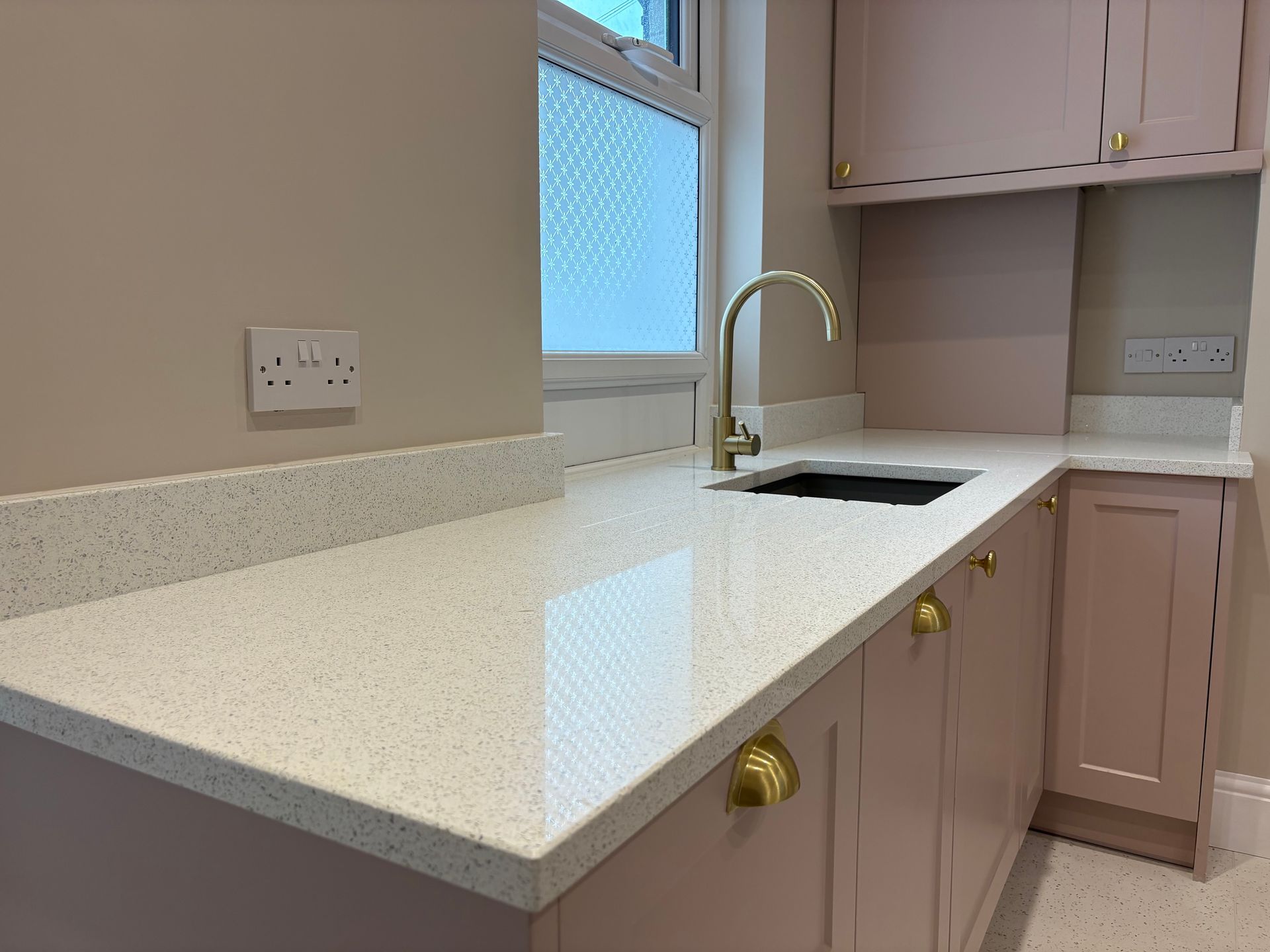 Pink kitchen cabinets with a white countertop, gold faucet, and window.