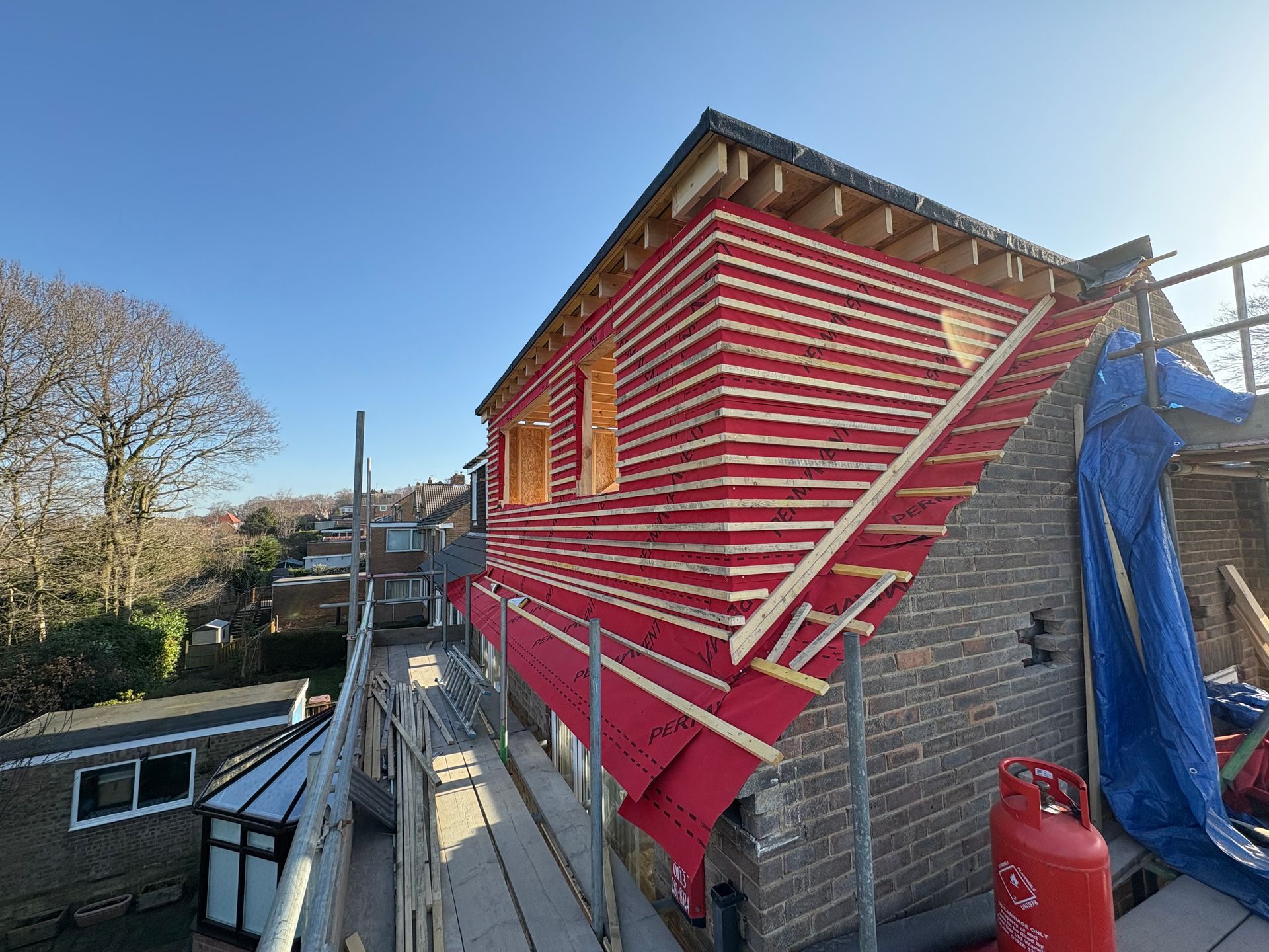 Construction on a building's exterior with red siding and scaffolding, under a clear sky.
