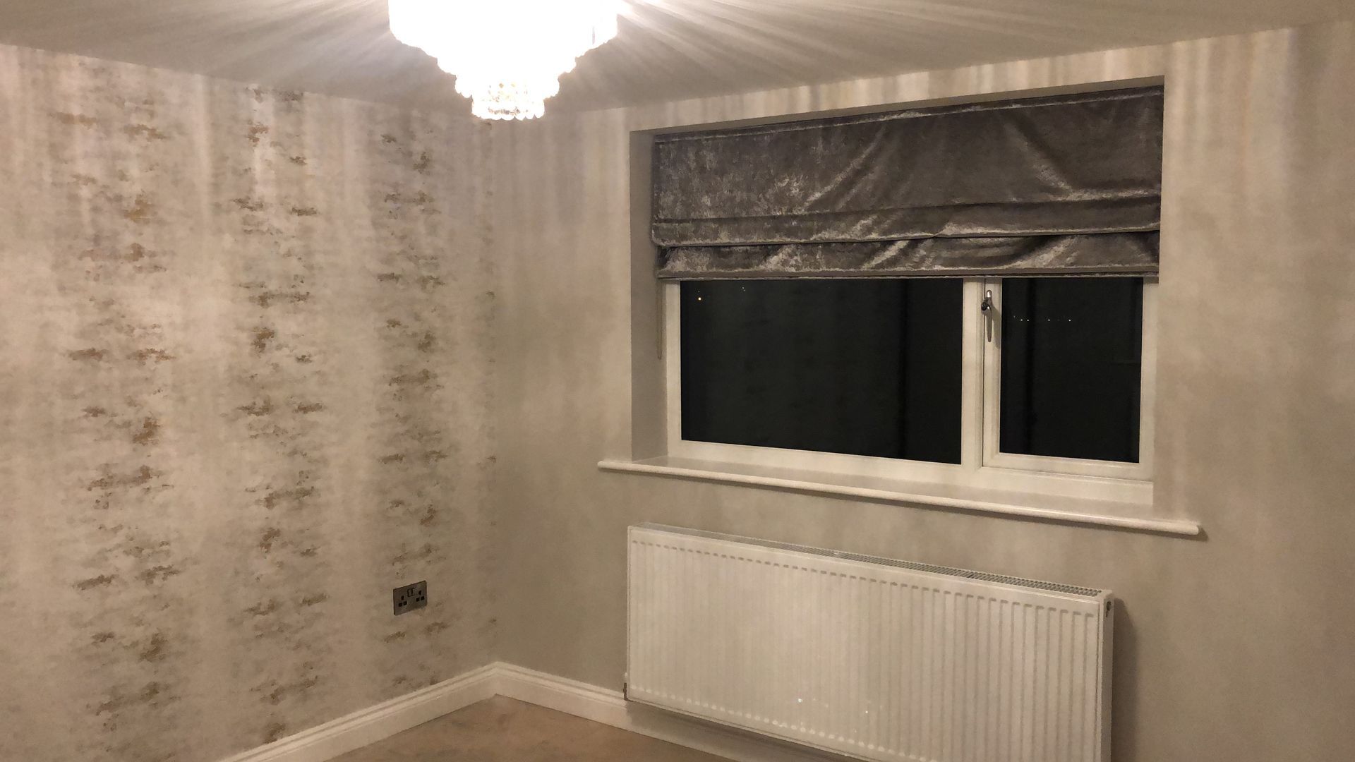 Room with patterned wallpaper, window with shade, radiator, and chandelier.
