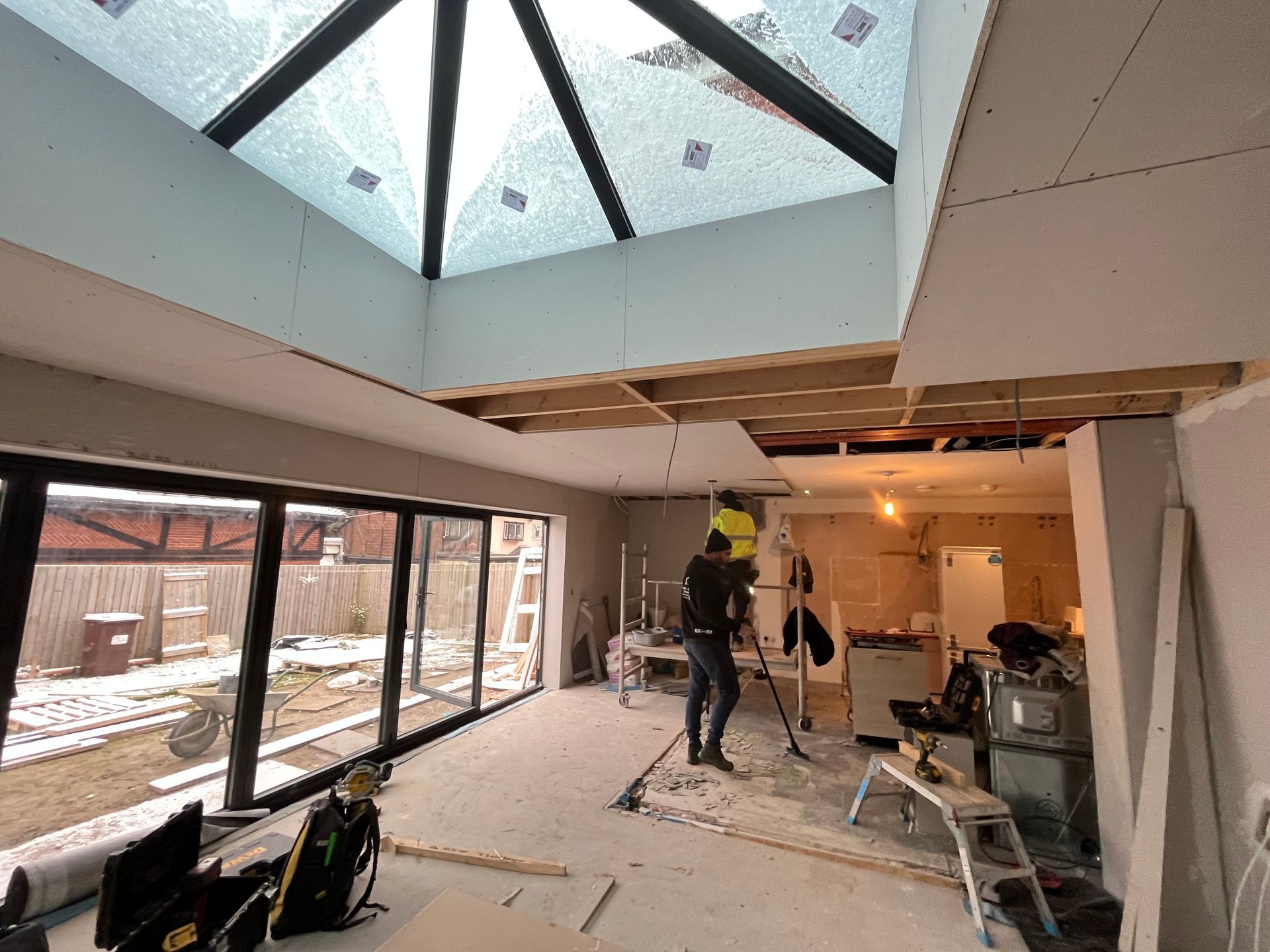 A room under construction with a skylight, a worker sweeping, and a view of the backyard.