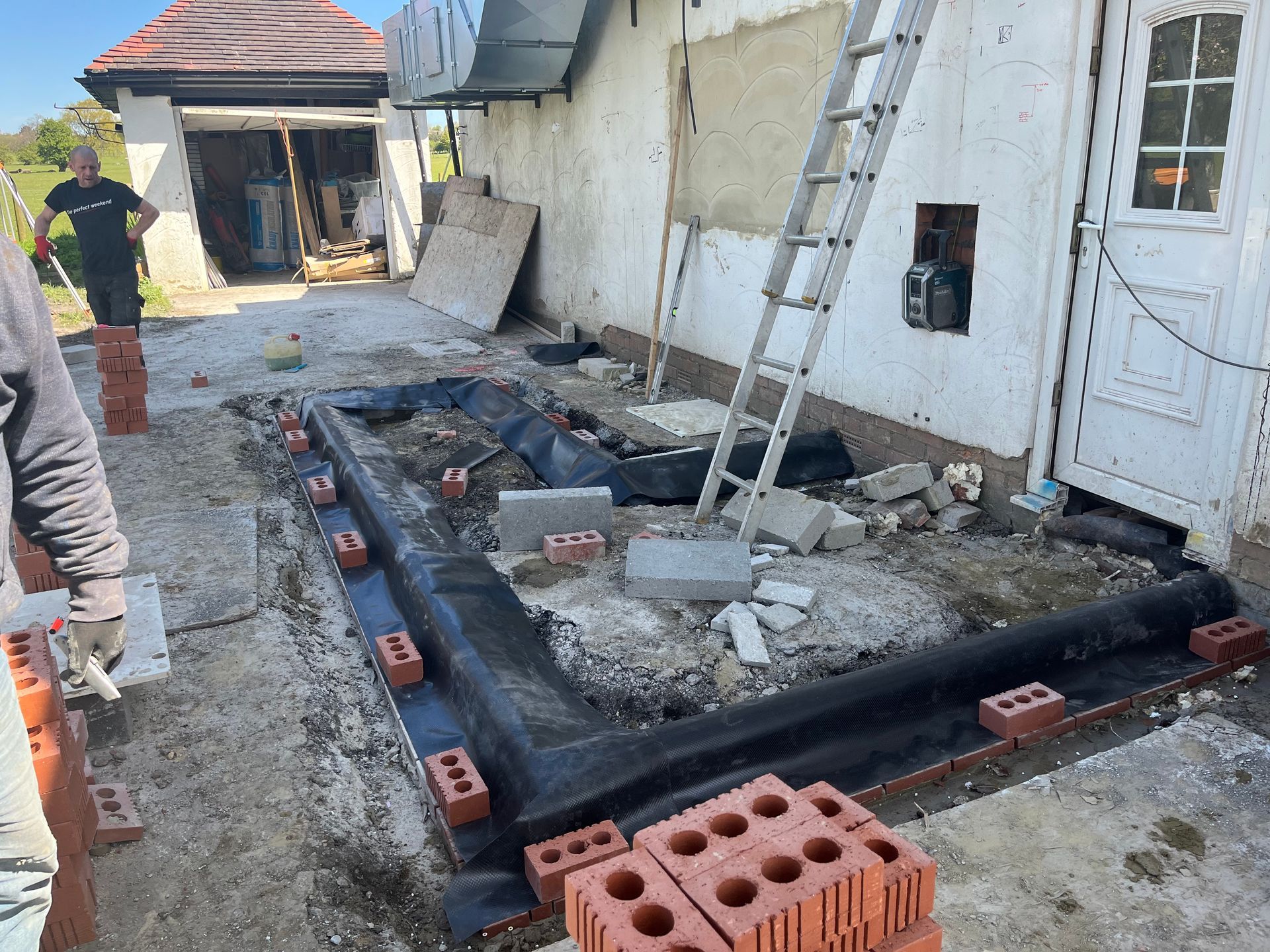 Construction site: bricks lining a dark membrane, near a building and garage. Person in background.