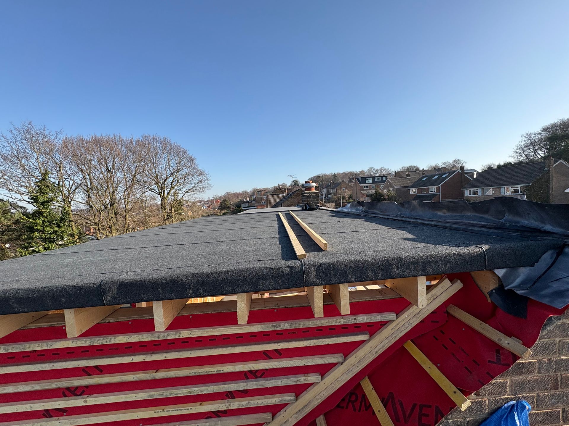 A flat roof under construction with black roofing material, wooden beams, and red sheathing