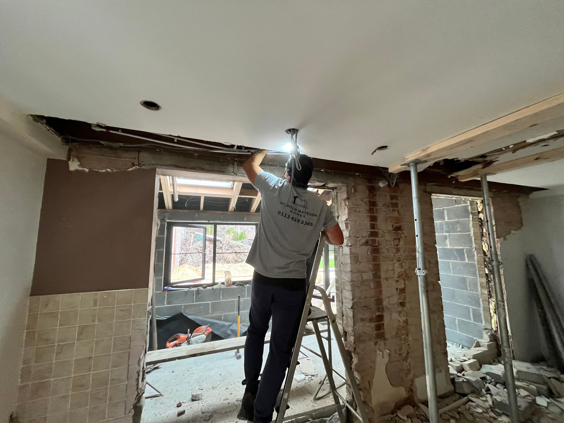 Man on a ladder working on ceiling demolition in a room with exposed brick and drywall.