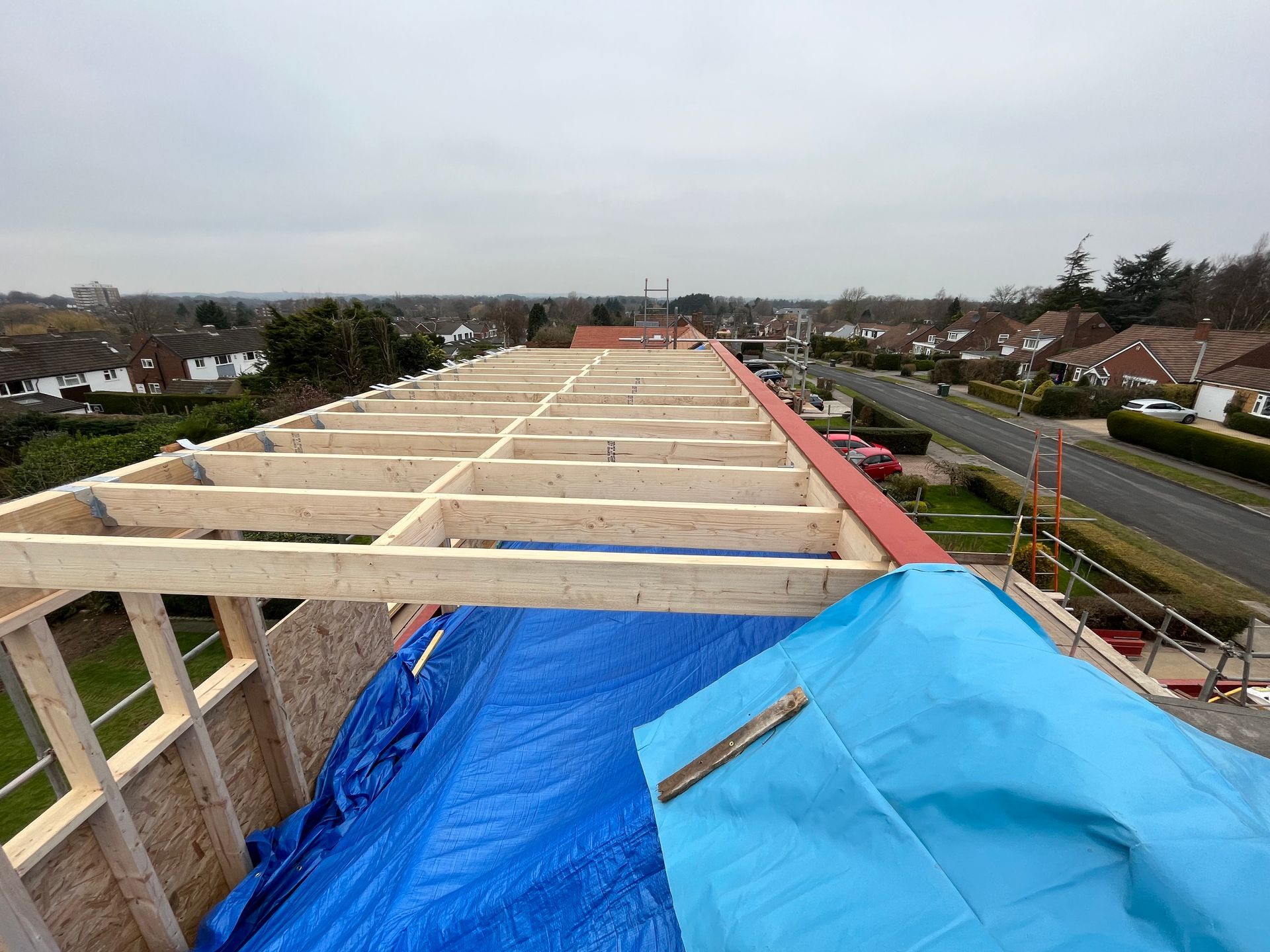 Wooden roof frame under construction with blue tarp and suburban background.