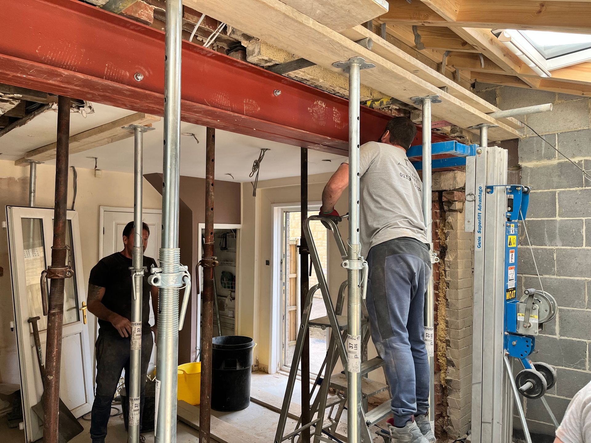 Two construction workers installing a red steel beam in a building. Scaffolding and supports are in place.