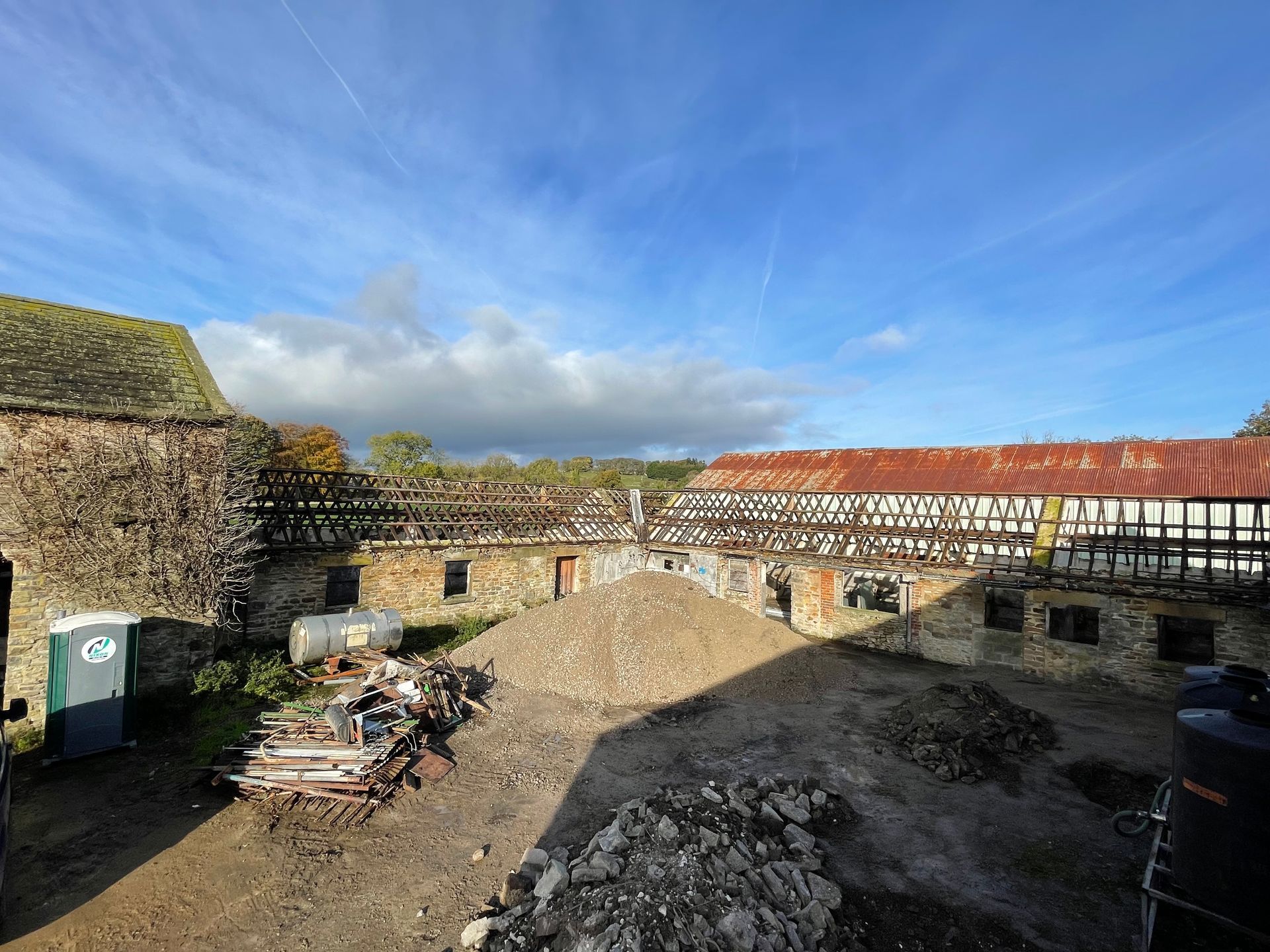 Barn courtyard under construction; piles of rubble and exposed beams under blue sky.