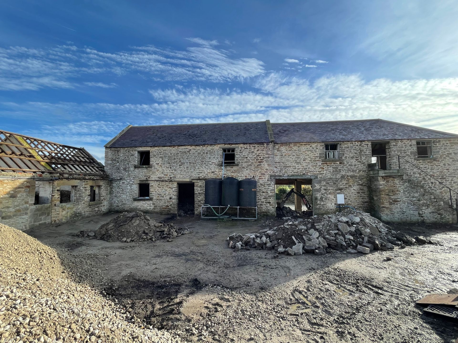 Stone barn under construction; exterior view on a sunny day, with exposed beams and debris.