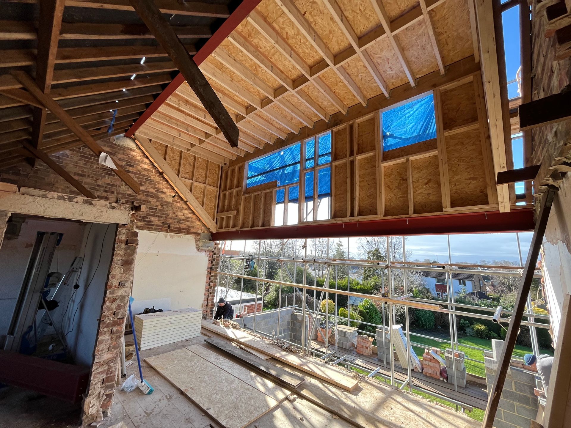 Construction site with exposed framing, red beam, windows, and blue tarp, with a view of the outdoors.