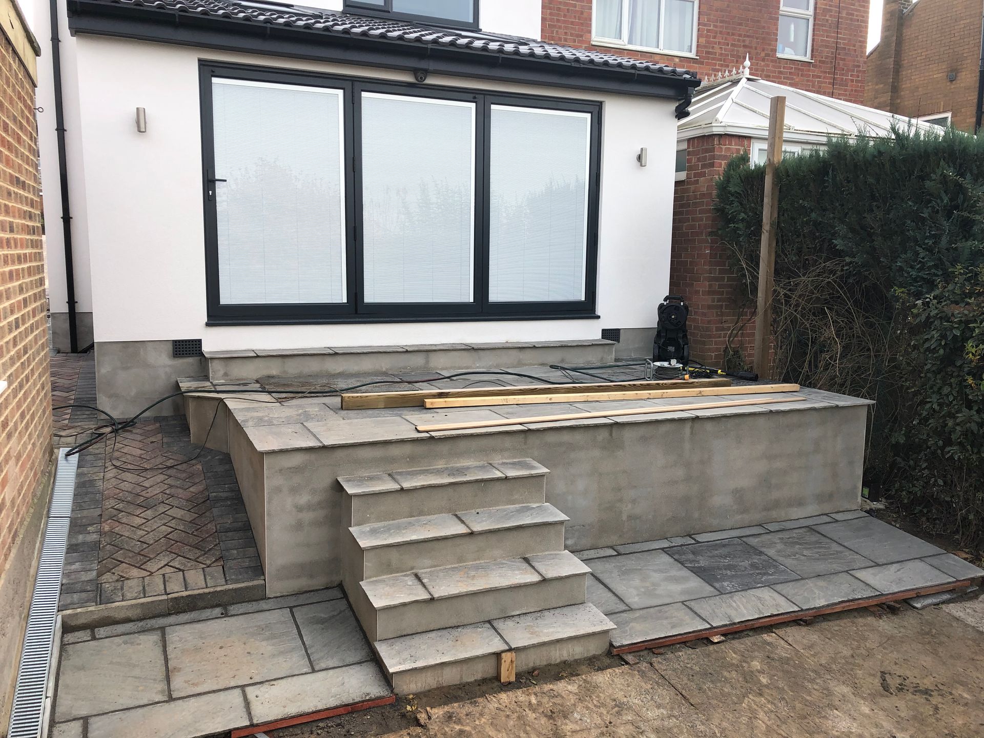 A patio under construction with gray stone steps and a concrete retaining wall, next to a house.