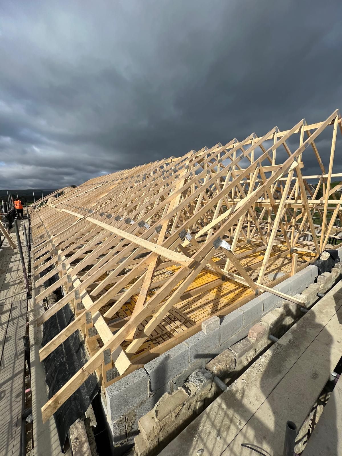 Wooden roof trusses being built on a brick structure with a cloudy sky overhead.