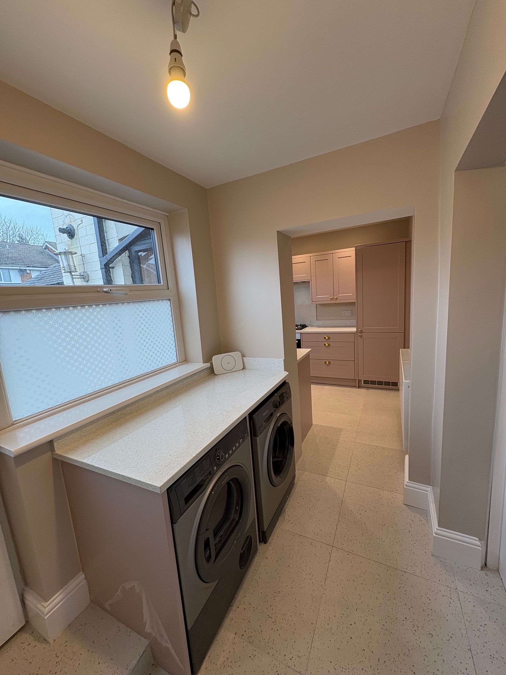 A laundry room with washing machine, dryer, counter, and doorway into a kitchen. Pink cabinets and beige walls.