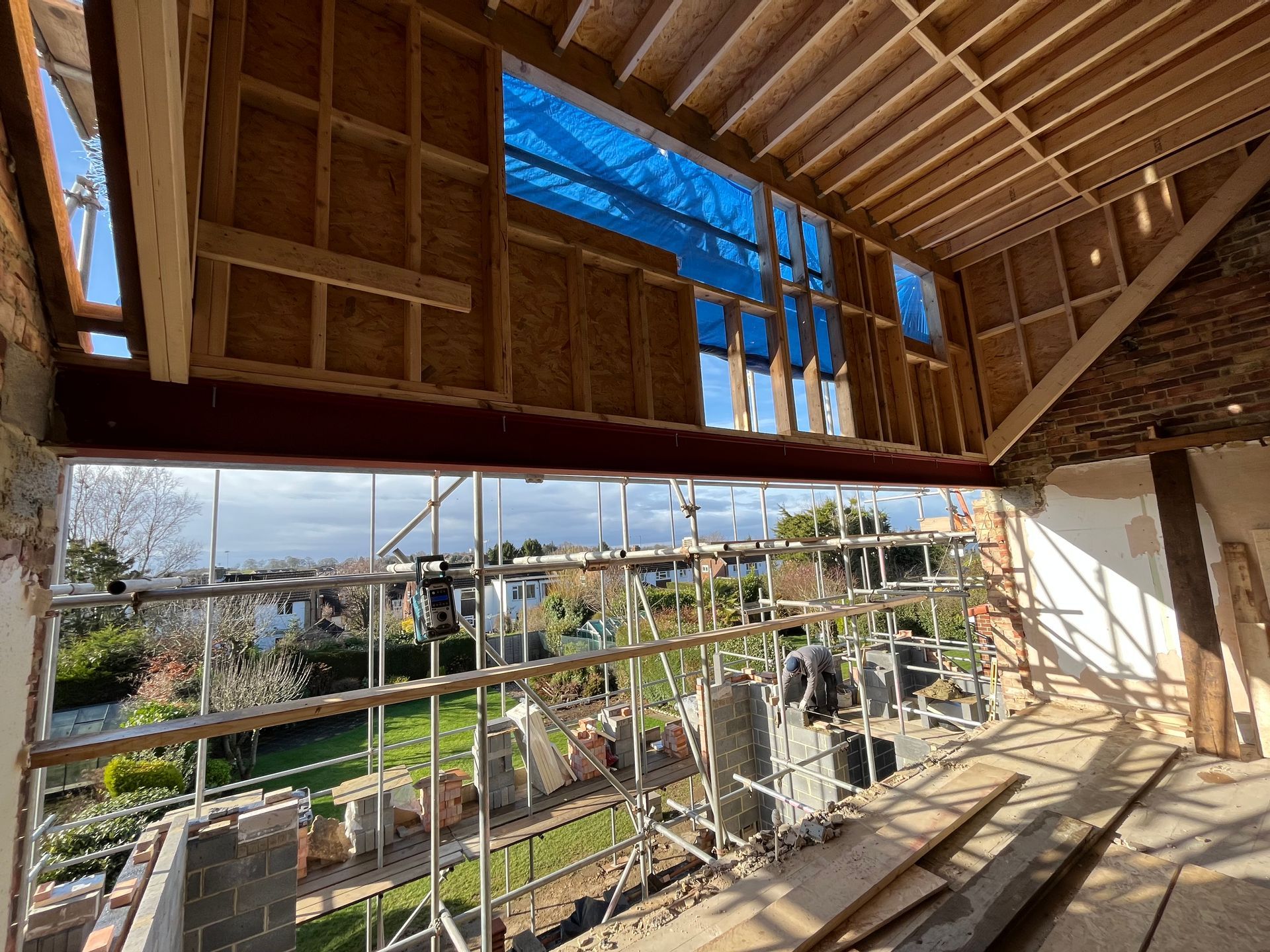 Interior view of a house under construction, showing exposed beams, scaffolding, and a view of the outside.