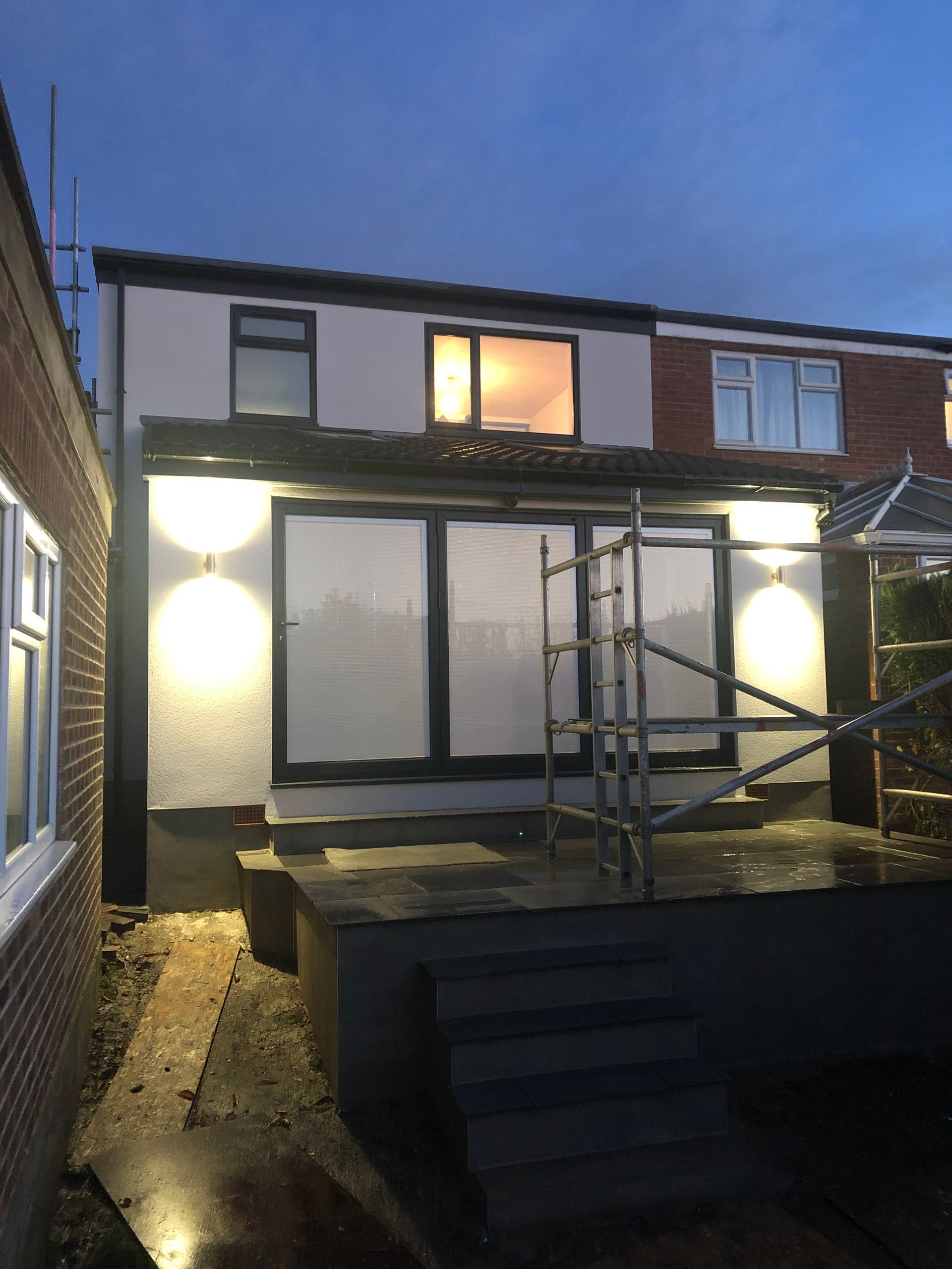 Back of house with large windows, lights, and scaffolding against a dusk sky.