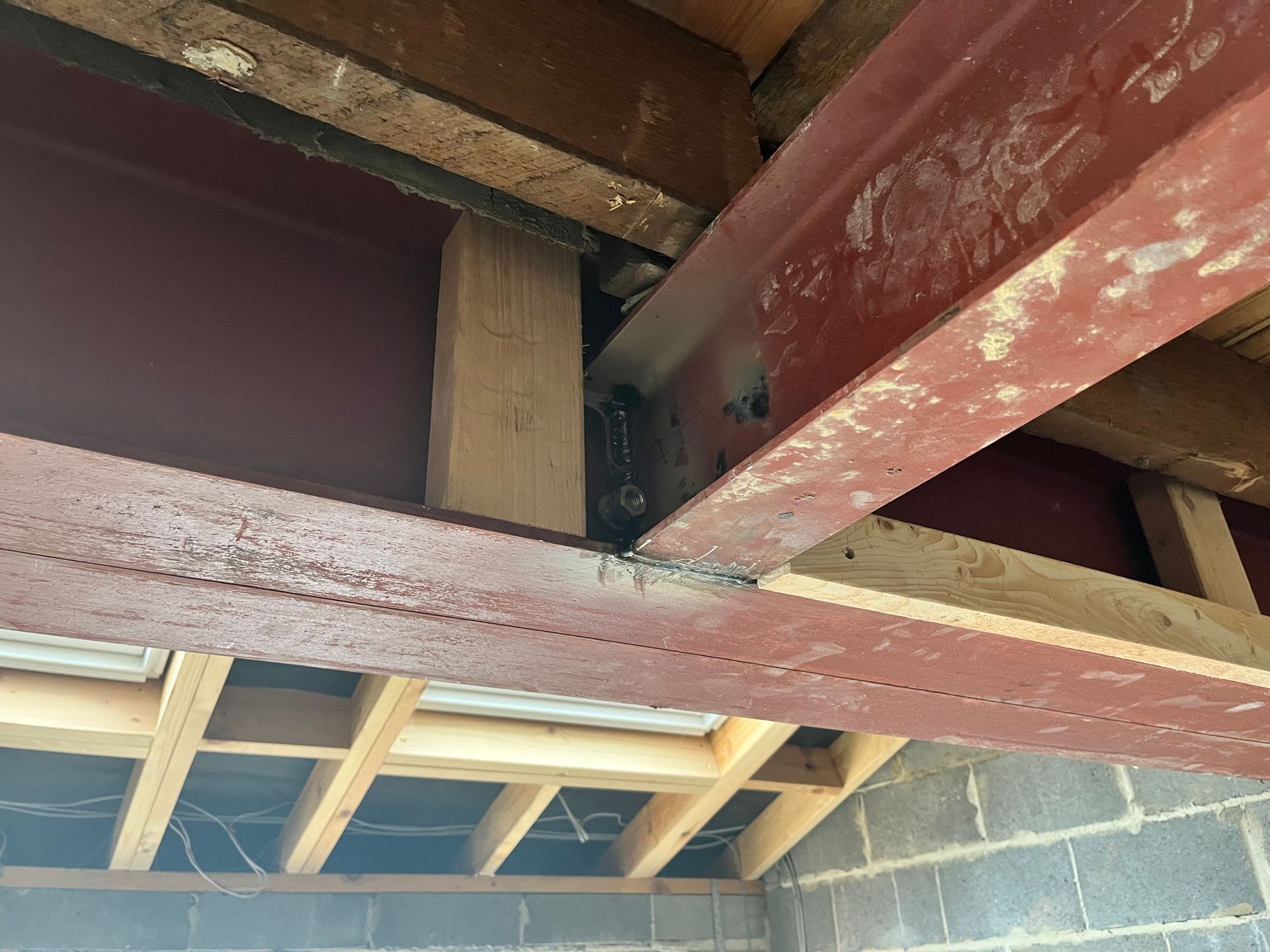 Red steel beams support a ceiling with wooden joists. A wooden support post is visible. Brick wall in bottom corner.