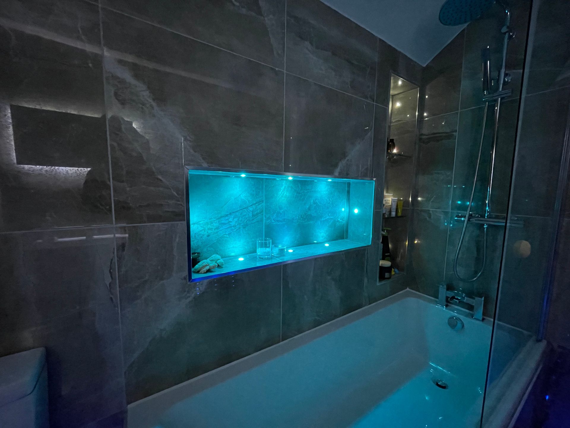 Bathroom with tub, built-in shelf with blue lights, and gray marble tiles.
