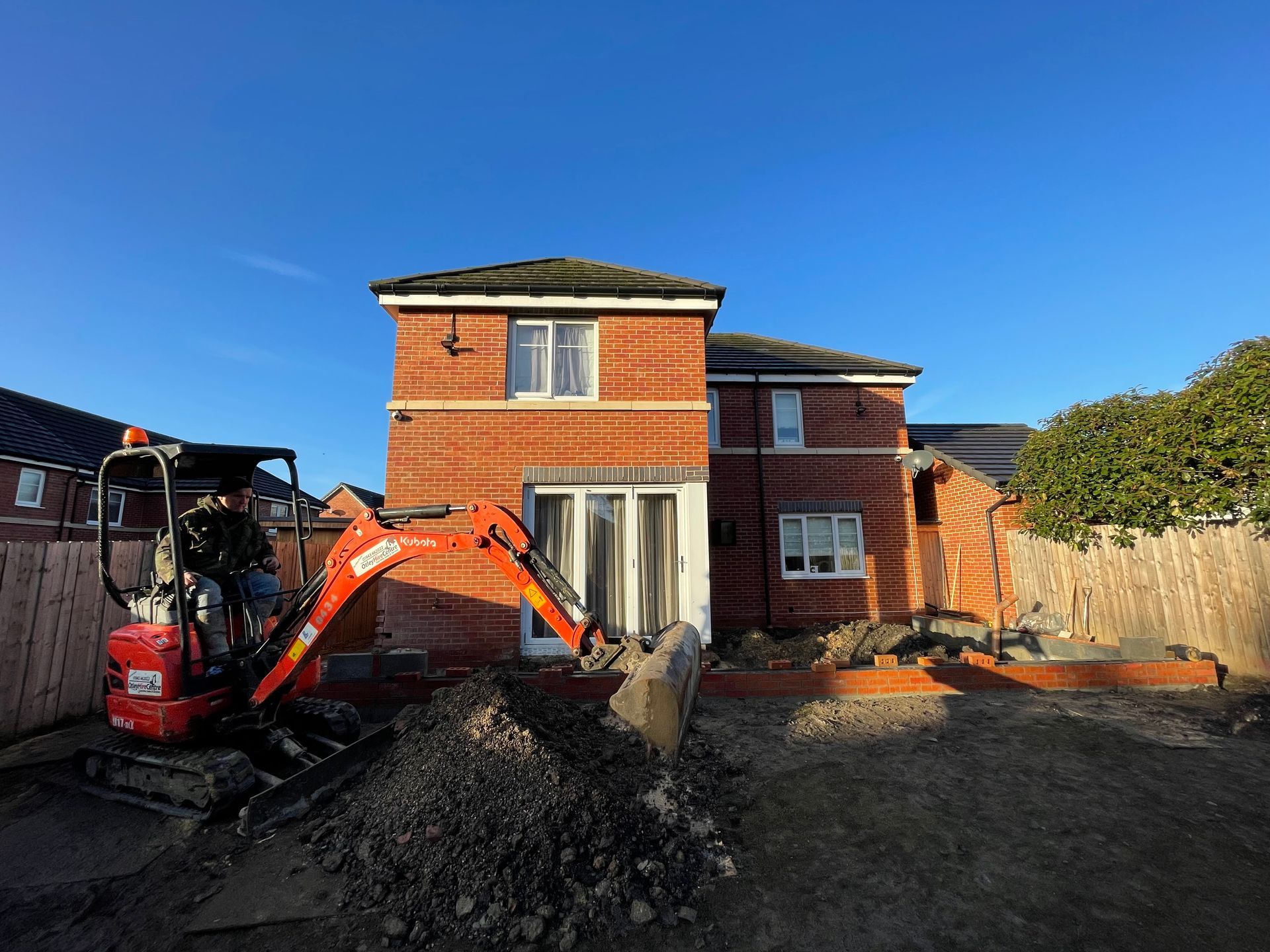 Construction: Mini excavator in front of a brick house, digging in the yard. Sunny day.
