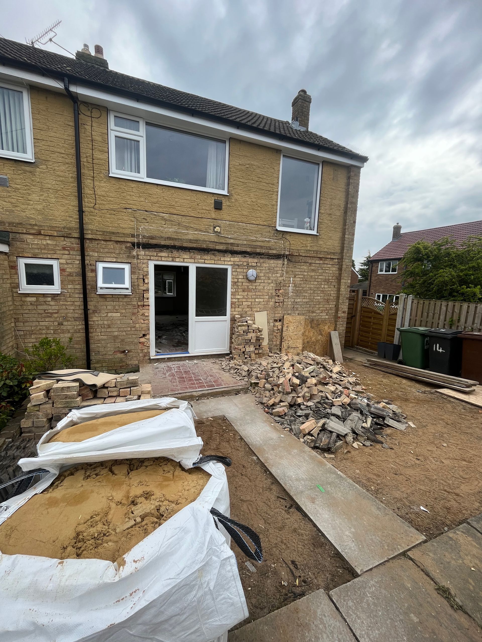 Backyard undergoing construction; bricks, sandbags, and a concrete path. Beige house with windows.