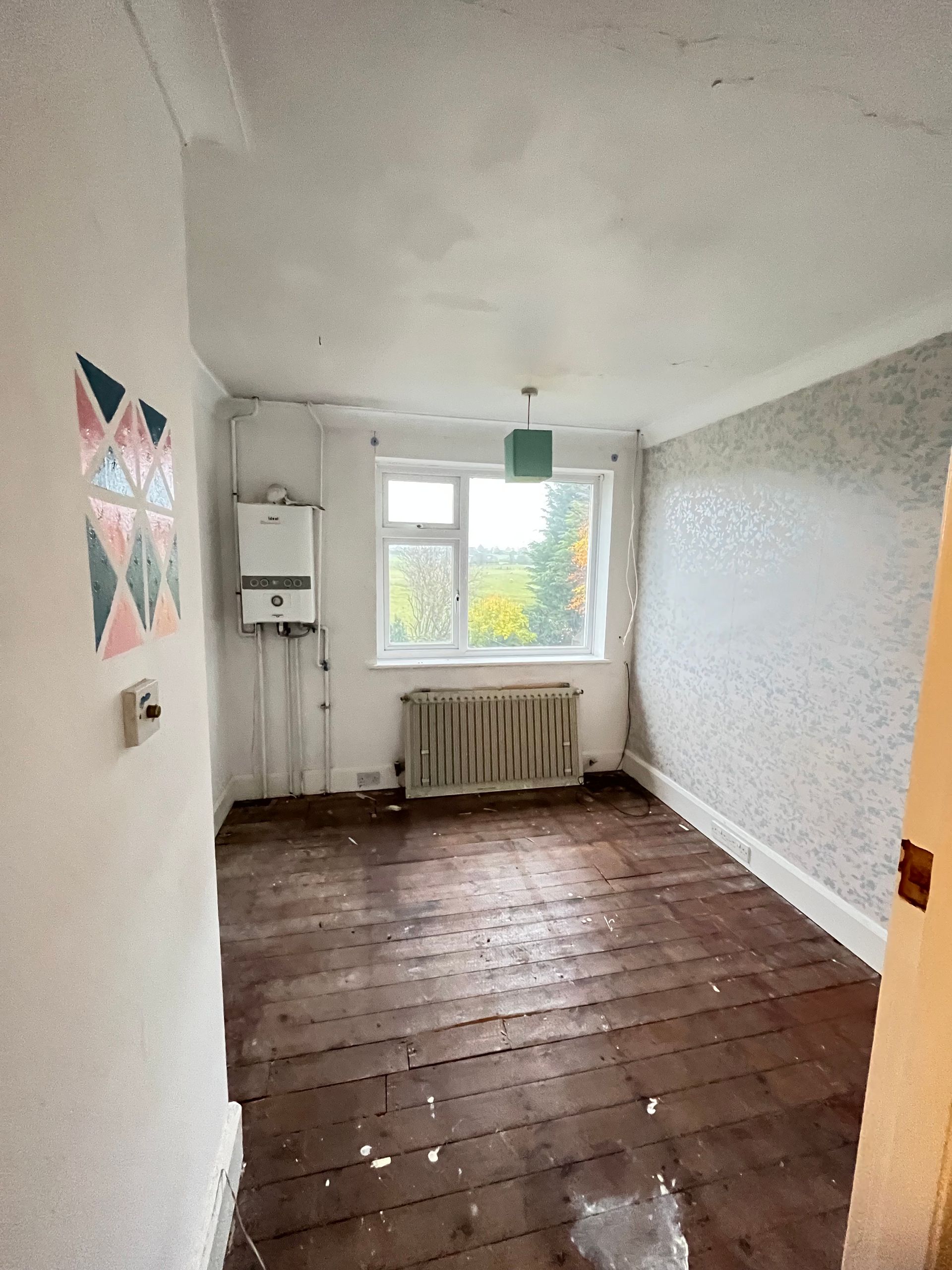 Interior view of a room with a gas boiler, window, radiator, and damaged wooden floor.