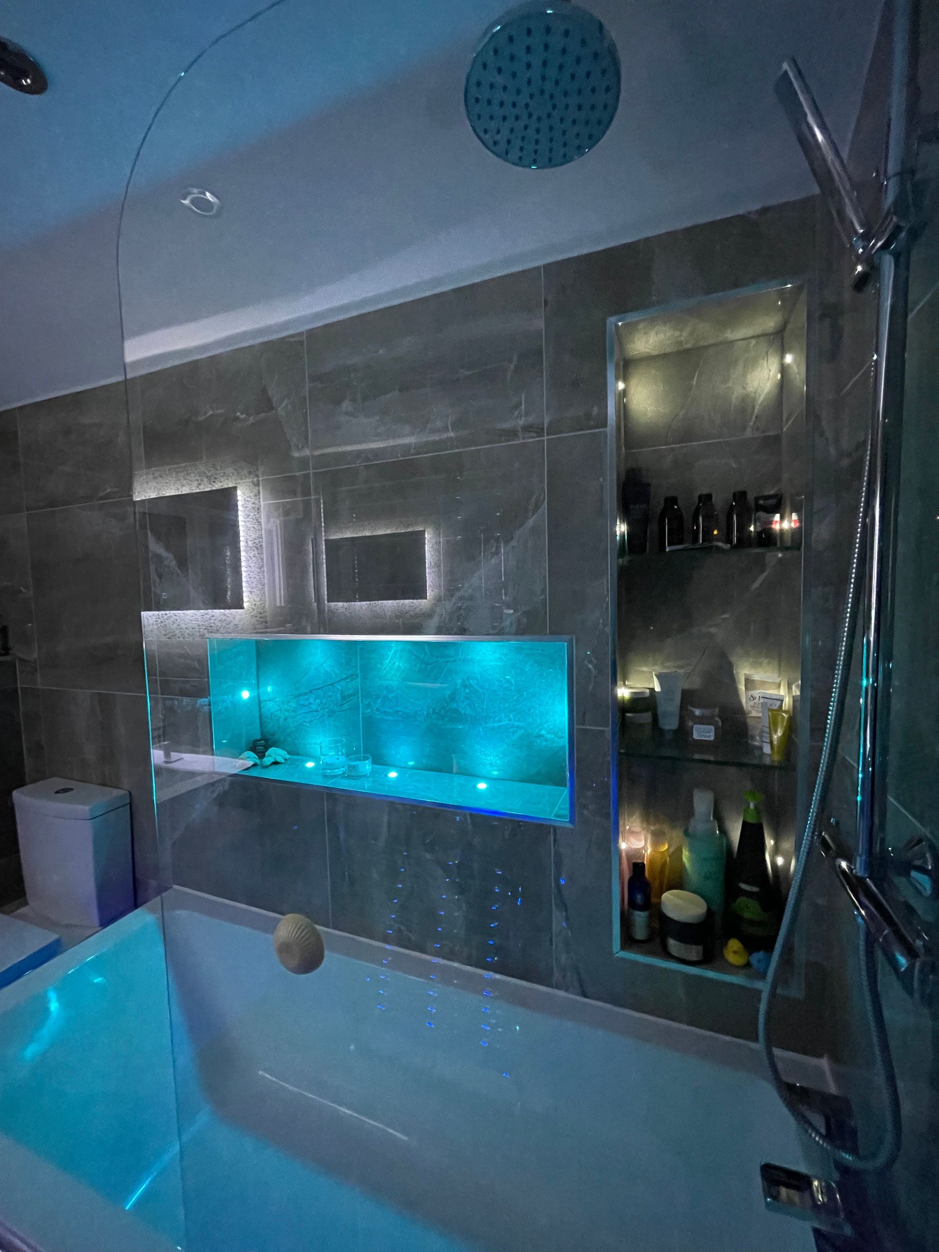 Modern bathroom with built-in shelves, blue LED lighting, and a white bathtub. Grey tiled walls.