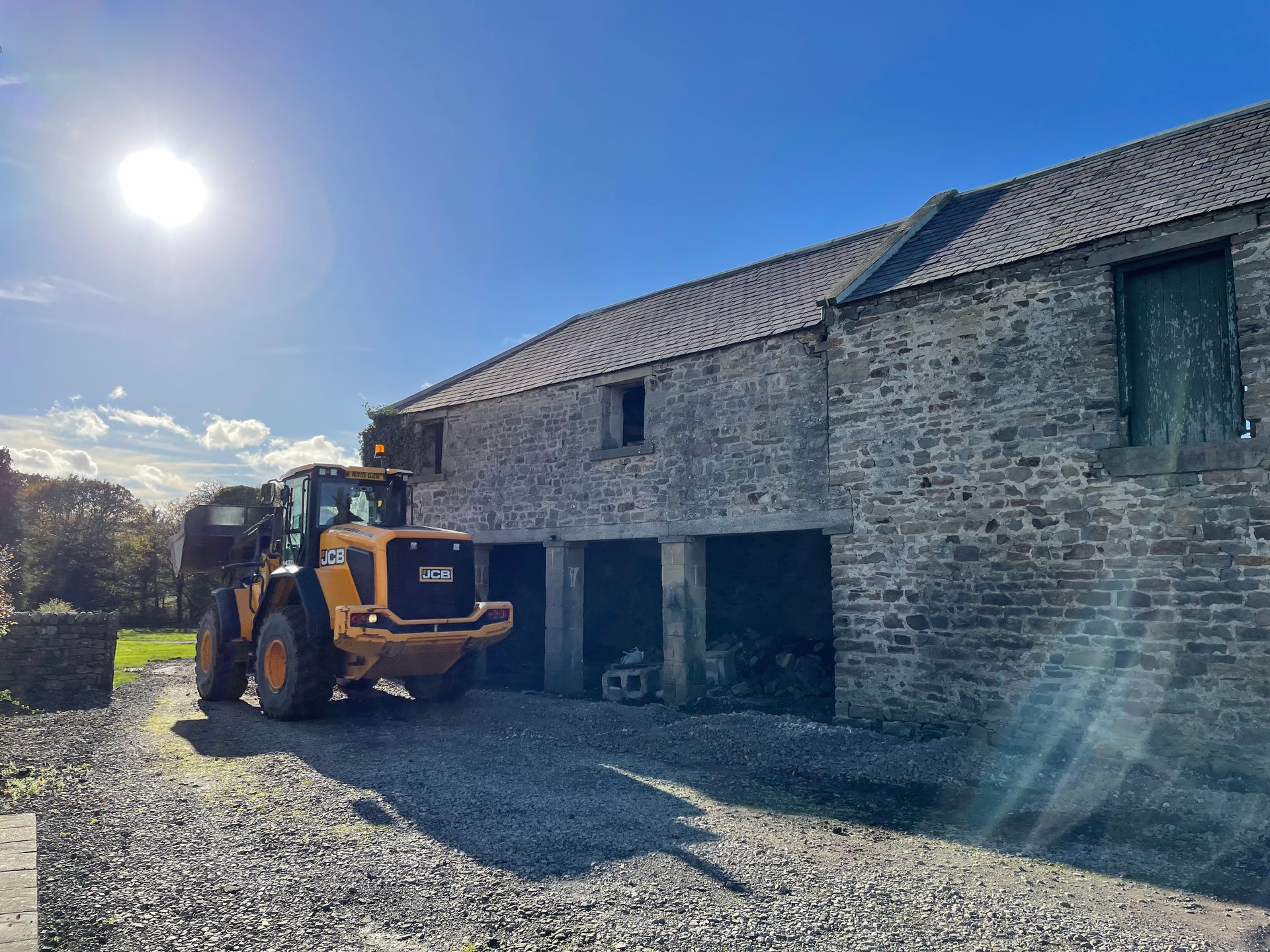 Yellow loader by a stone barn under a bright sunny sky.