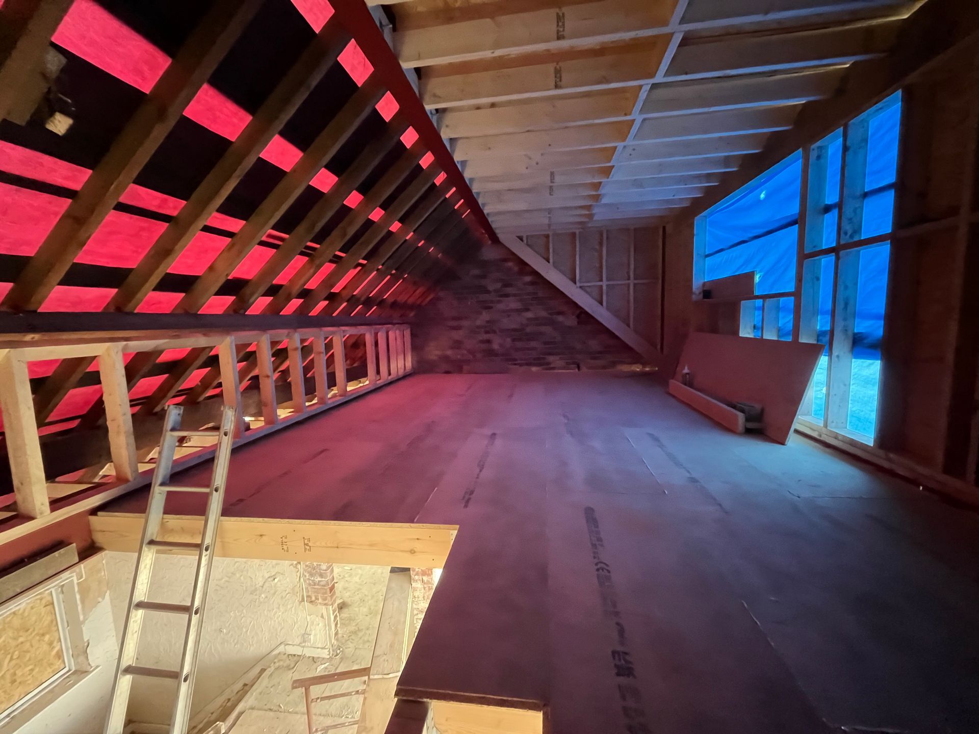 Interior of a partially constructed attic space with wood framing and red sheeting; a ladder leans against a wall.