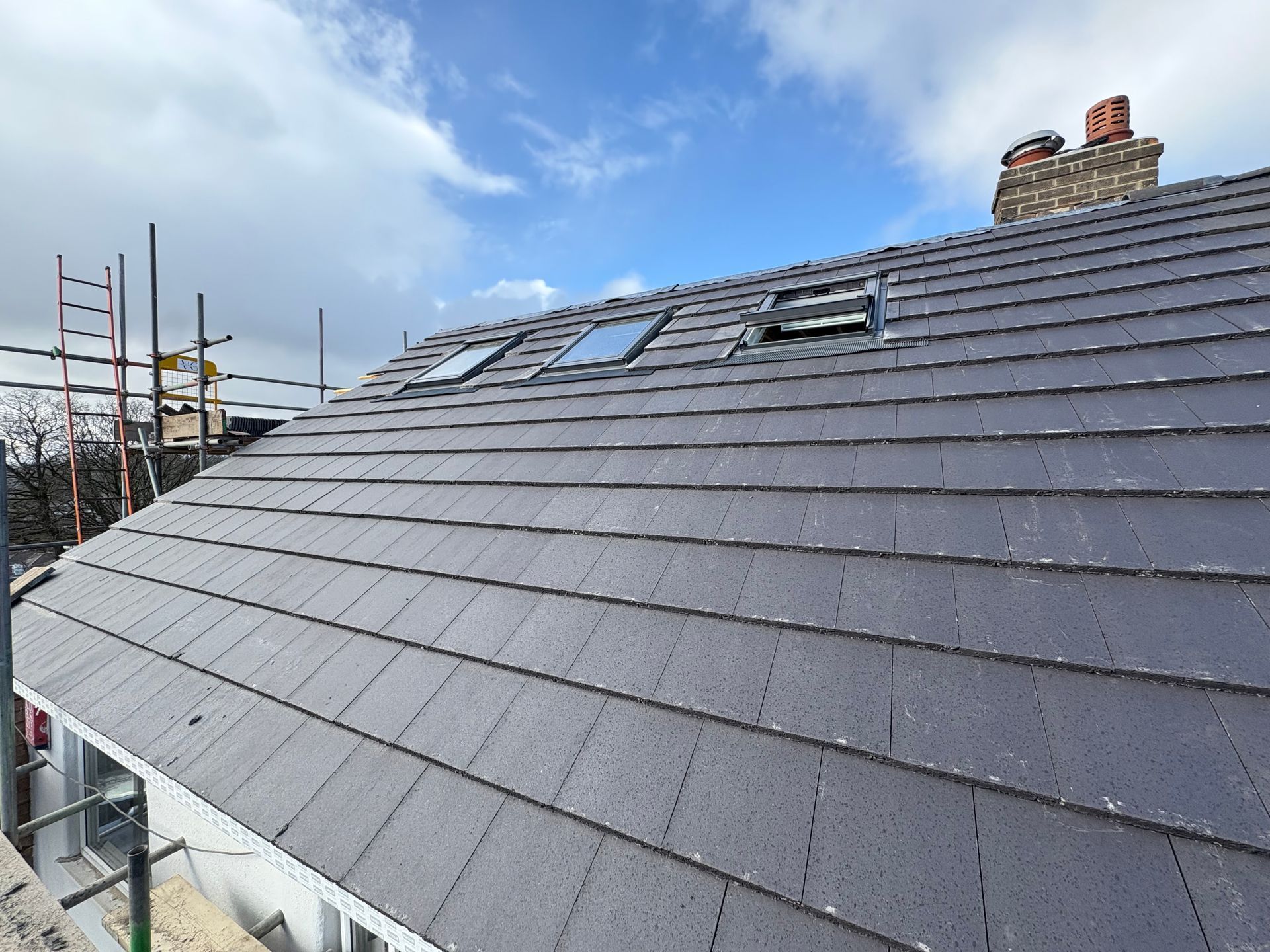 Slate roof with three skylights under a cloudy sky, scaffolding on left, chimney on right.