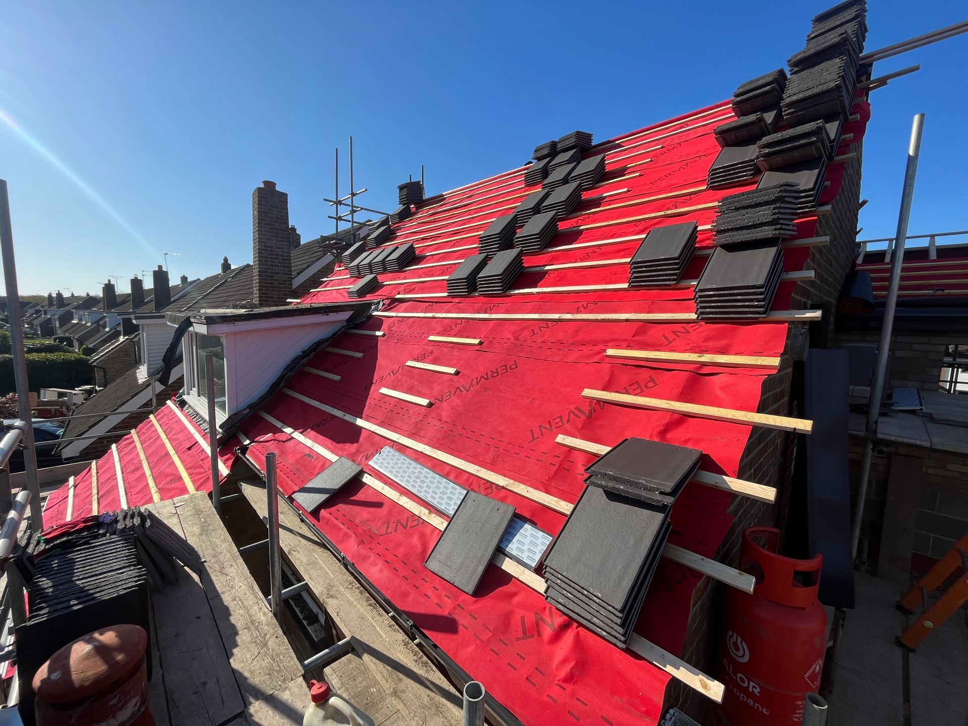 Roof under construction; red underlayment, black tiles stacked, scaffolding, clear blue sky.