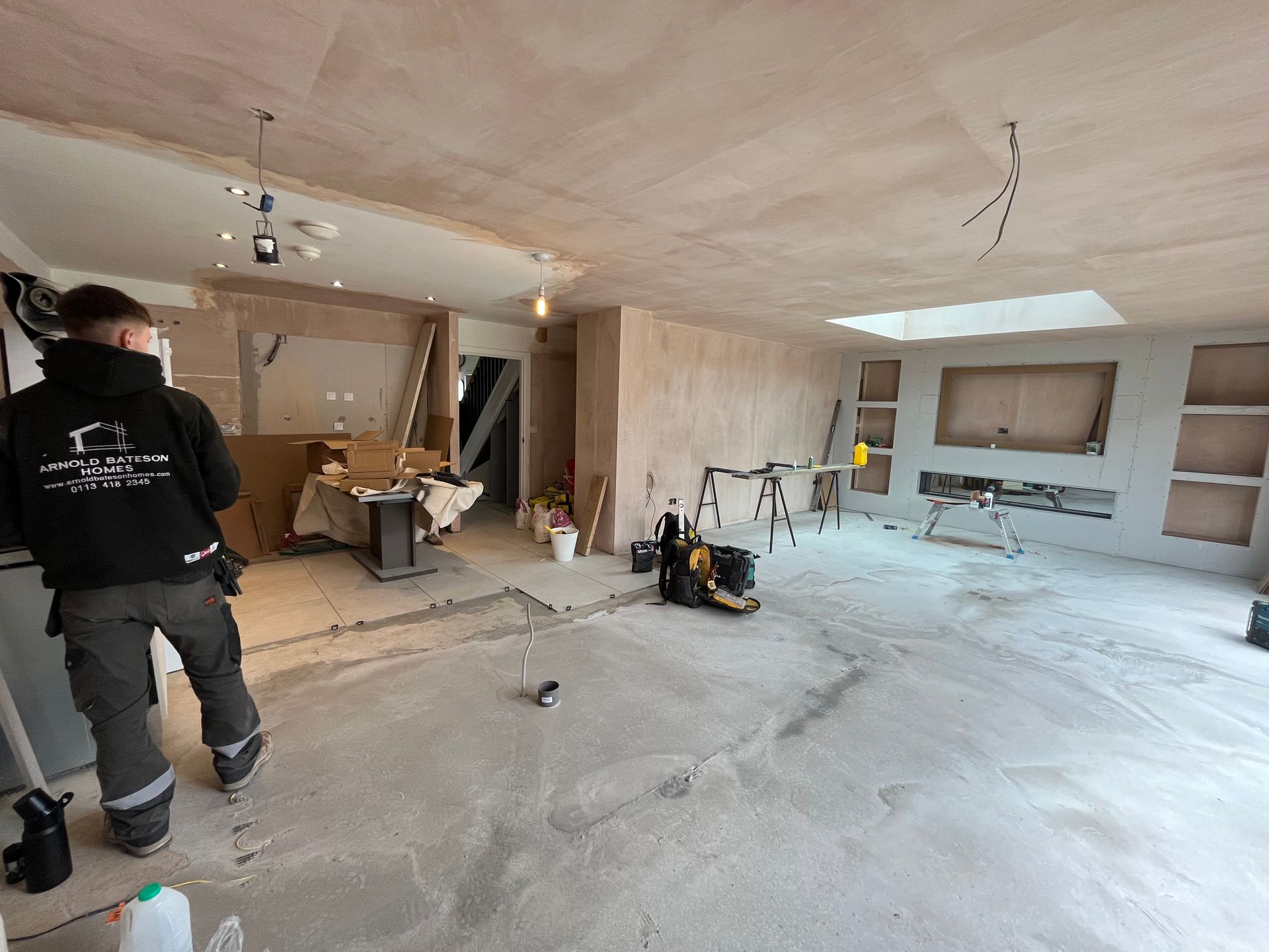 Person in work clothes in a room under construction; exposed wiring, concrete floor, freshly plastered walls.