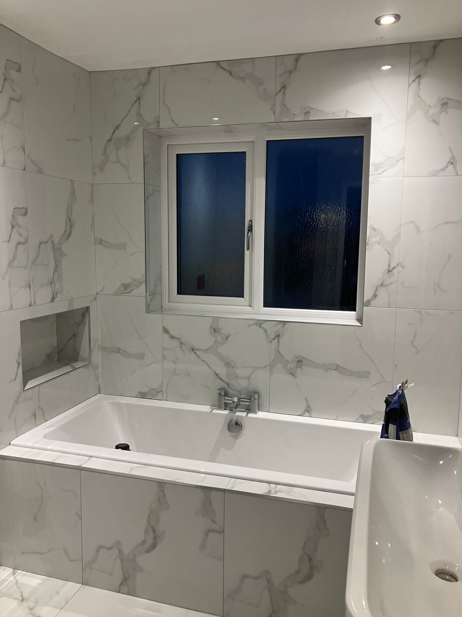 Bathroom with white marble tiles, a bathtub, window, and built-in shelf.