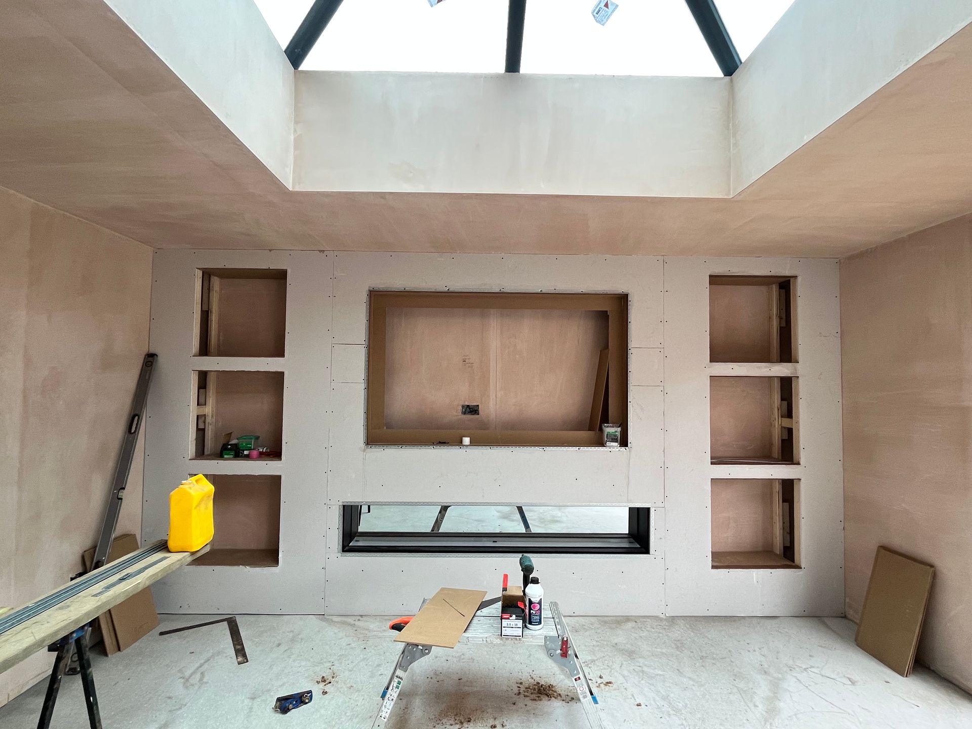 Interior room with a built-in fireplace, TV niche, and shelving under construction; natural light from skylight.