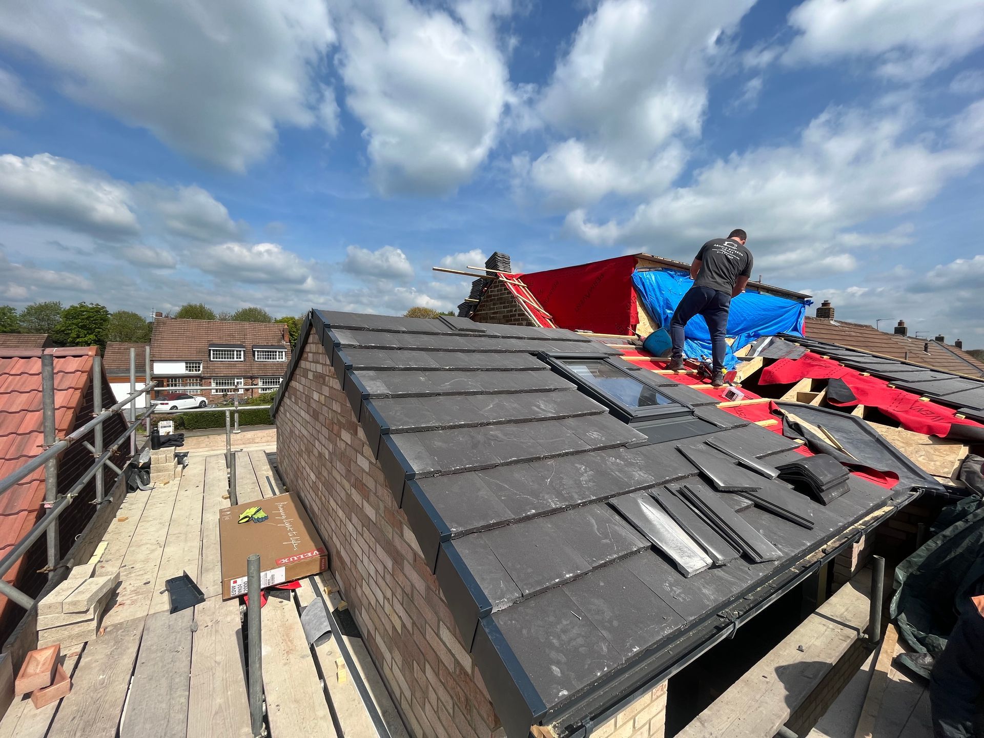 Roofer on a rooftop with new tiles and a partially tarped section under a blue sky.