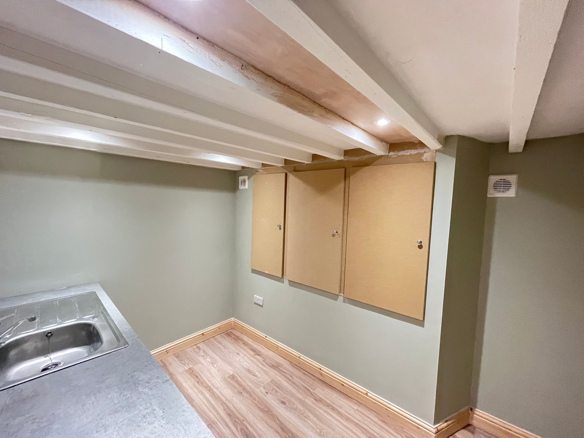 A small room with cabinets, sink, and exposed beams. Pale green walls, light wood floor, and a stainless steel sink.