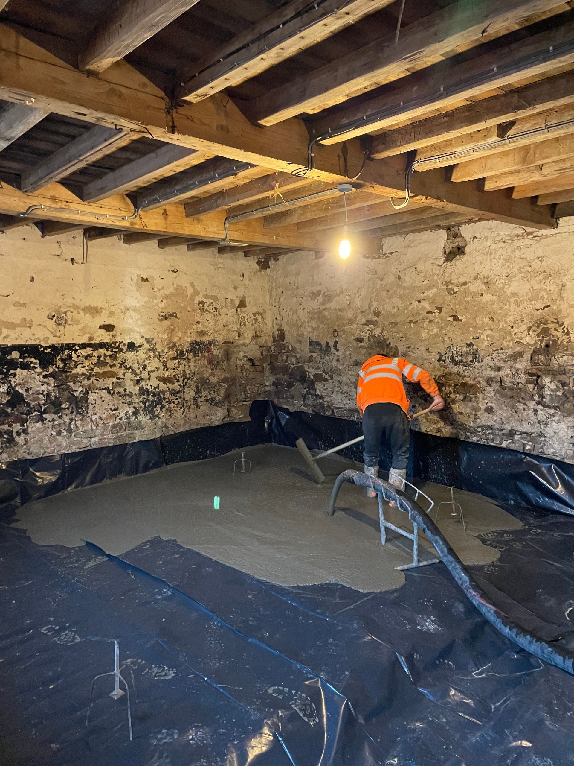 Man in orange vest leveling wet concrete floor in a room with exposed wooden beams, on black sheeting.