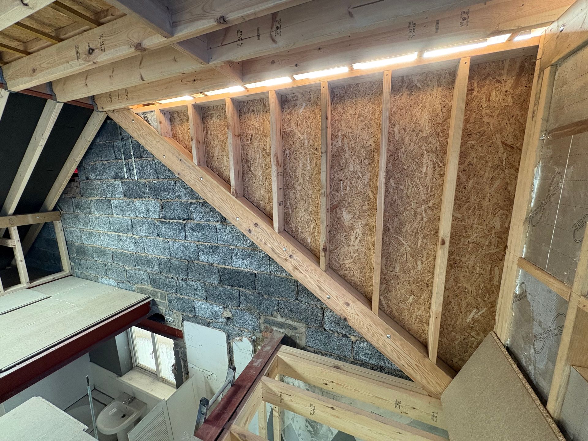 Interior construction of a sloped wall with wooden framework and OSB sheathing against a brick wall.