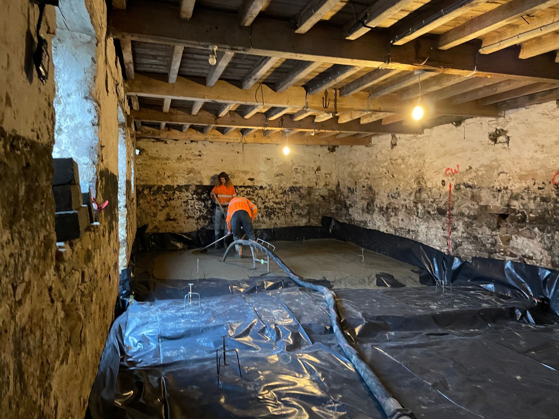 Two workers pouring concrete in a basement, plastic sheeting covers the floor, wood beams overhead.