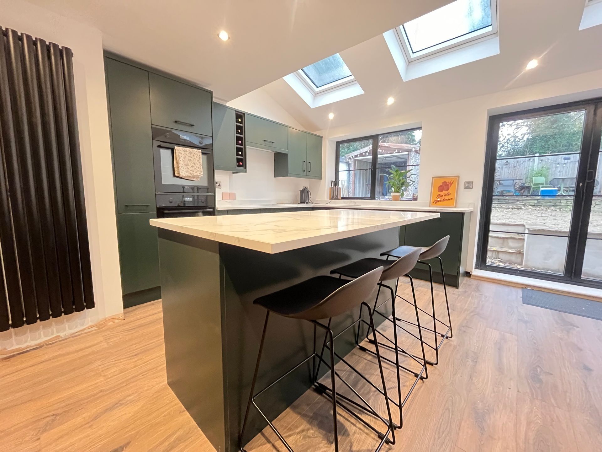 Modern kitchen with dark green cabinets, island with stools, skylights, and a view to the backyard.