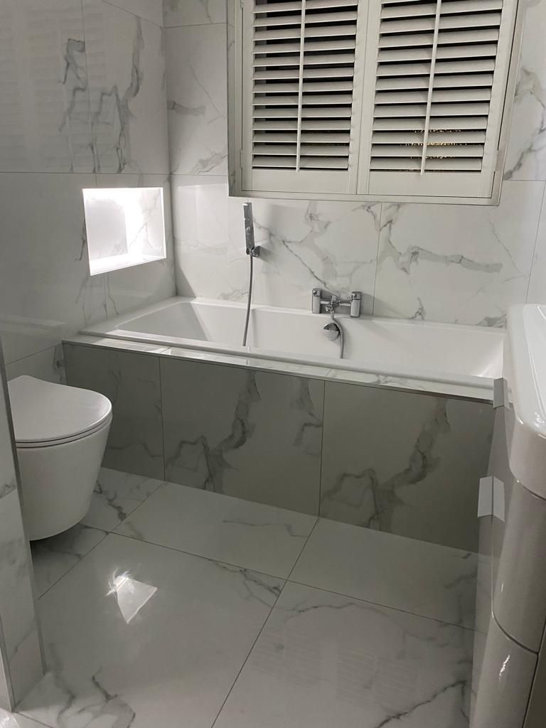 Modern bathroom with a white bathtub, toilet, and marble-patterned tiles; a window is visible above the tub.