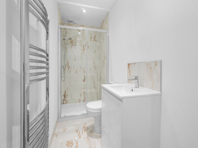 Small, white bathroom with marble shower and vanity. Chrome towel warmer.