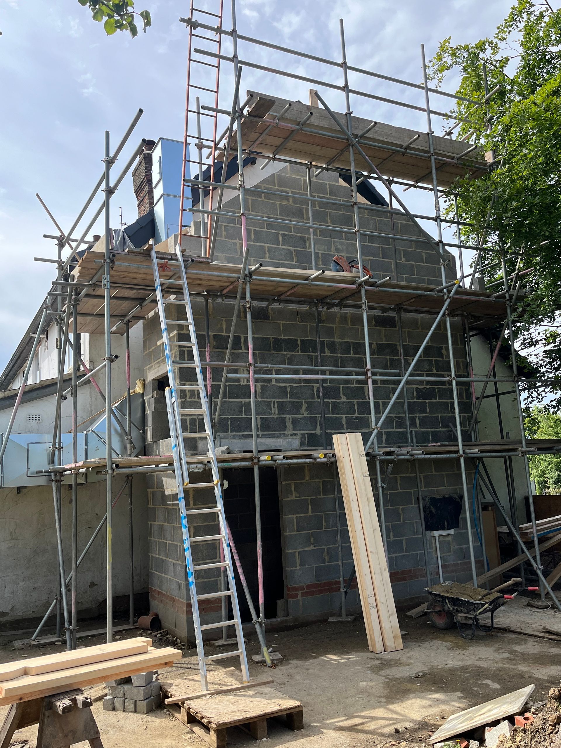 House under construction, scaffolding surrounding the grey block walls, ladder reaching the roof.