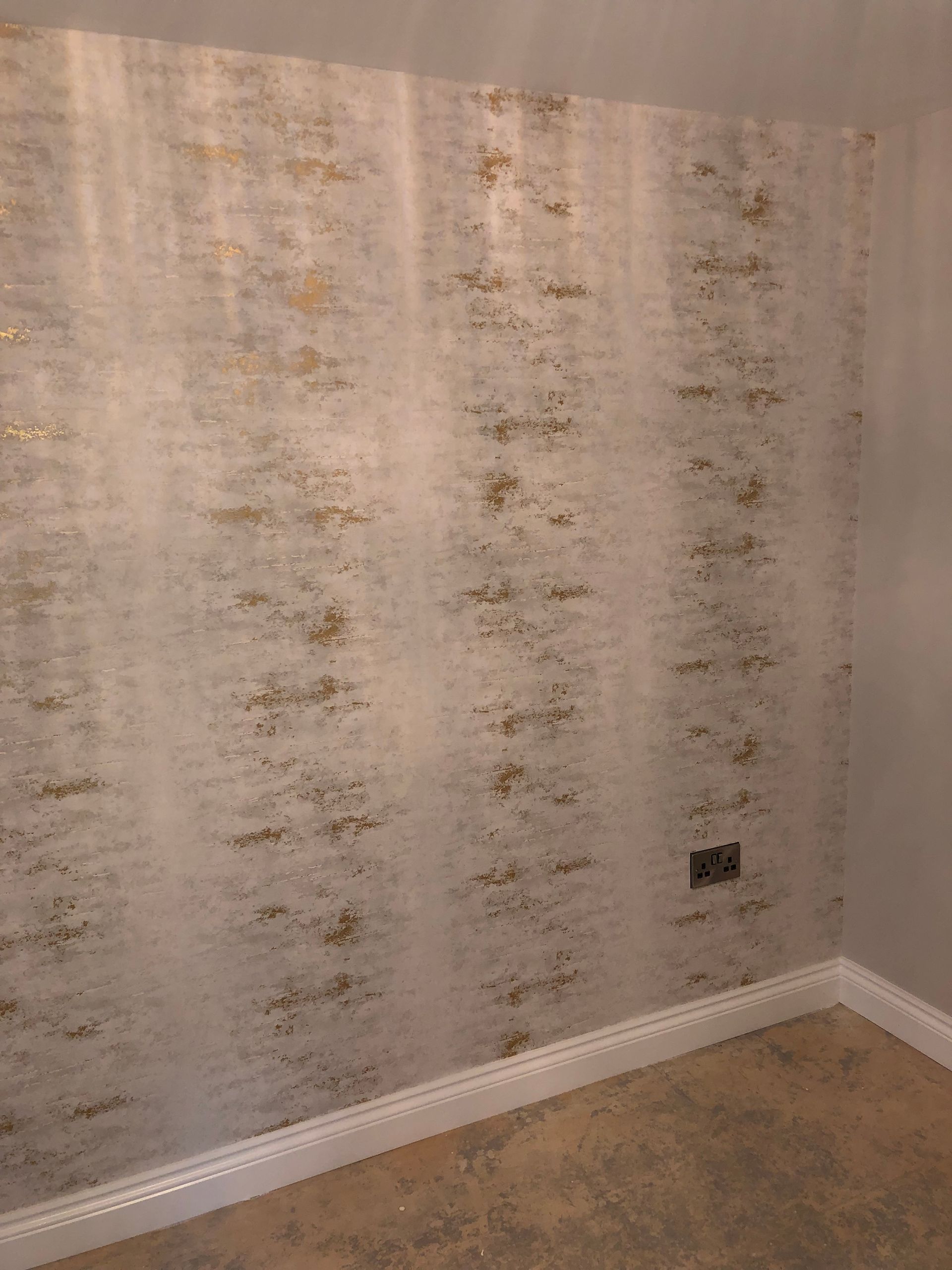 Wallpapered wall corner with light-colored textured design, gold accents, and a power outlet.