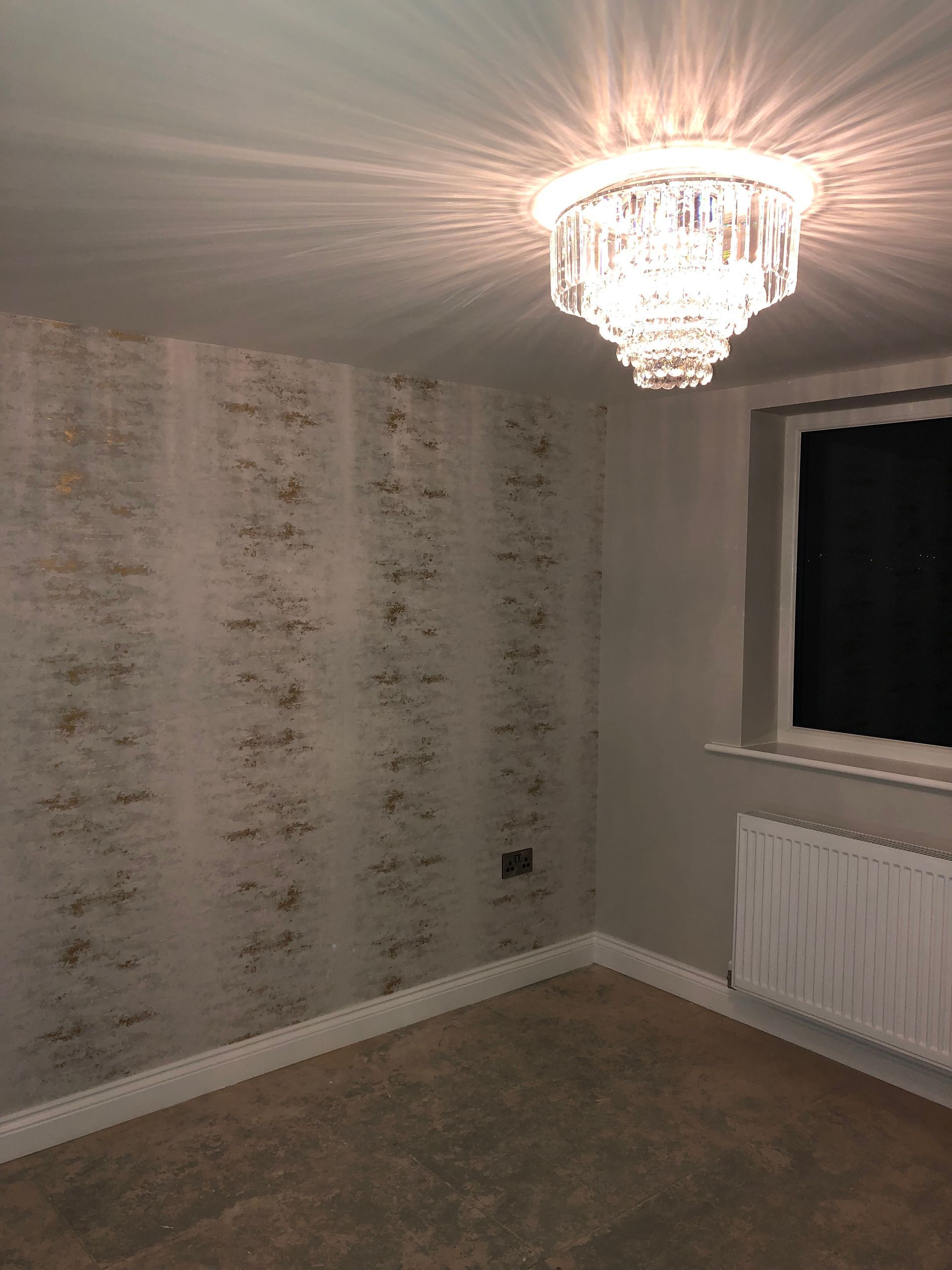 A room with patterned wallpaper, a chandelier, a window, and a radiator; lit by the chandelier.