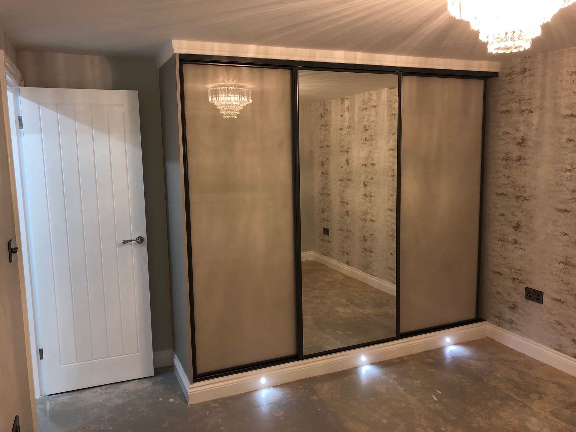 A modern bedroom with a three-panel sliding door wardrobe with mirrored center. A white door is on the left.