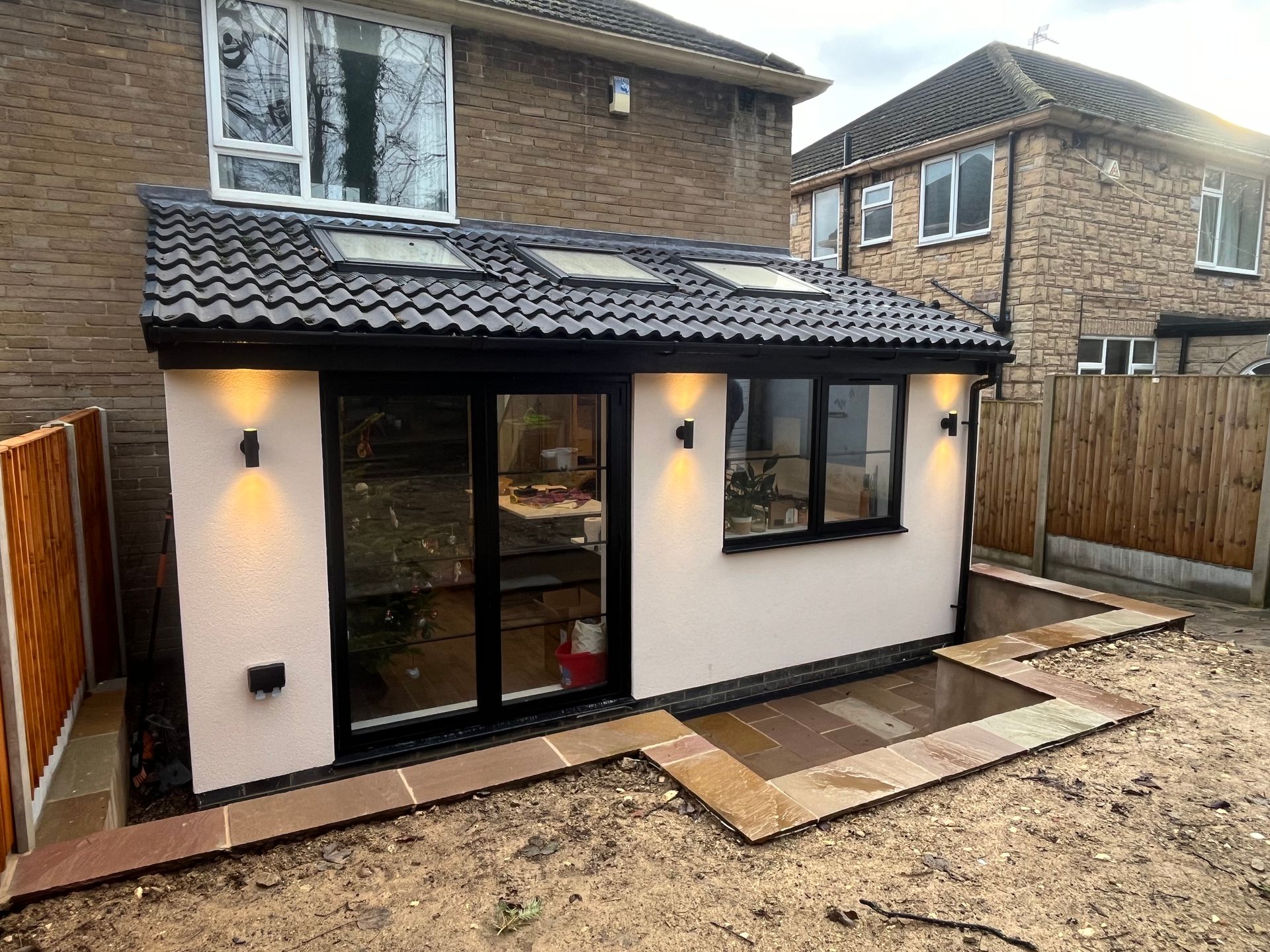 Extension with black-framed windows, skylights, and sconces. Pale pink walls, tiled roof, brick house in the background.