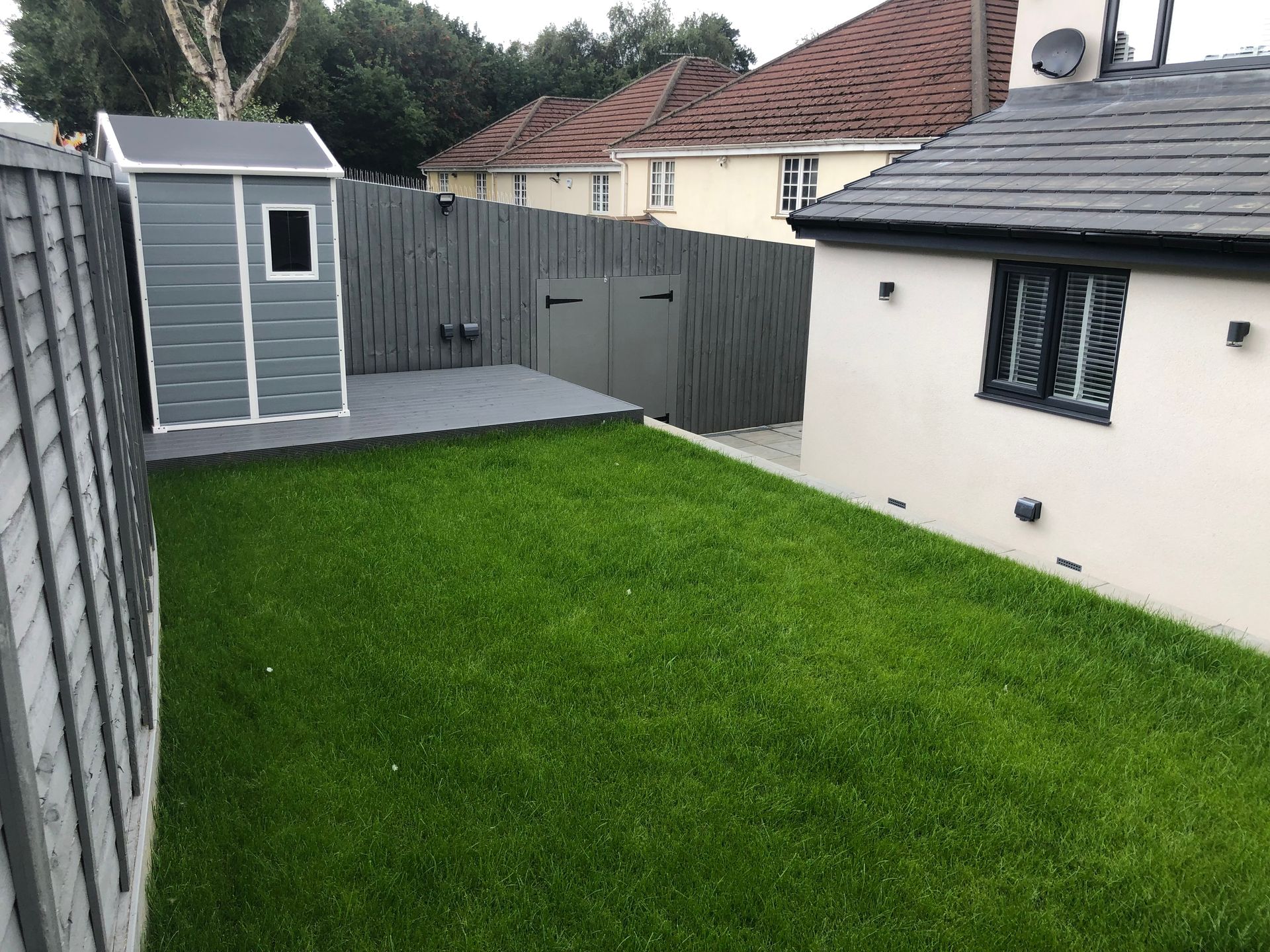 Lush green backyard with shed, deck, and house. Gray fencing and a white house with black trim.
